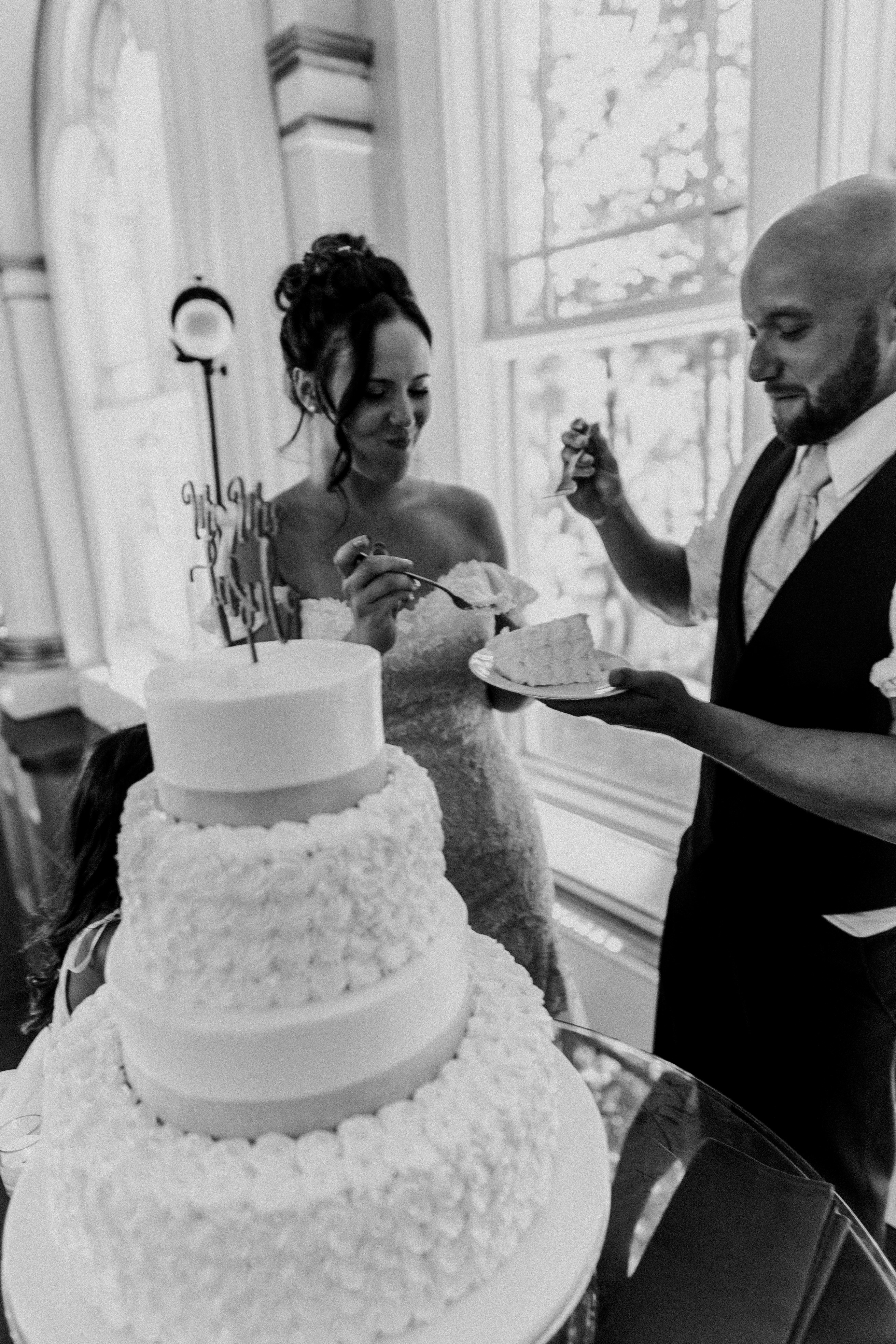 The couple cuts their three-tiered white wedding cake together near a bright window with sheer curtains. The bride wears a strapless white gown and has her hair