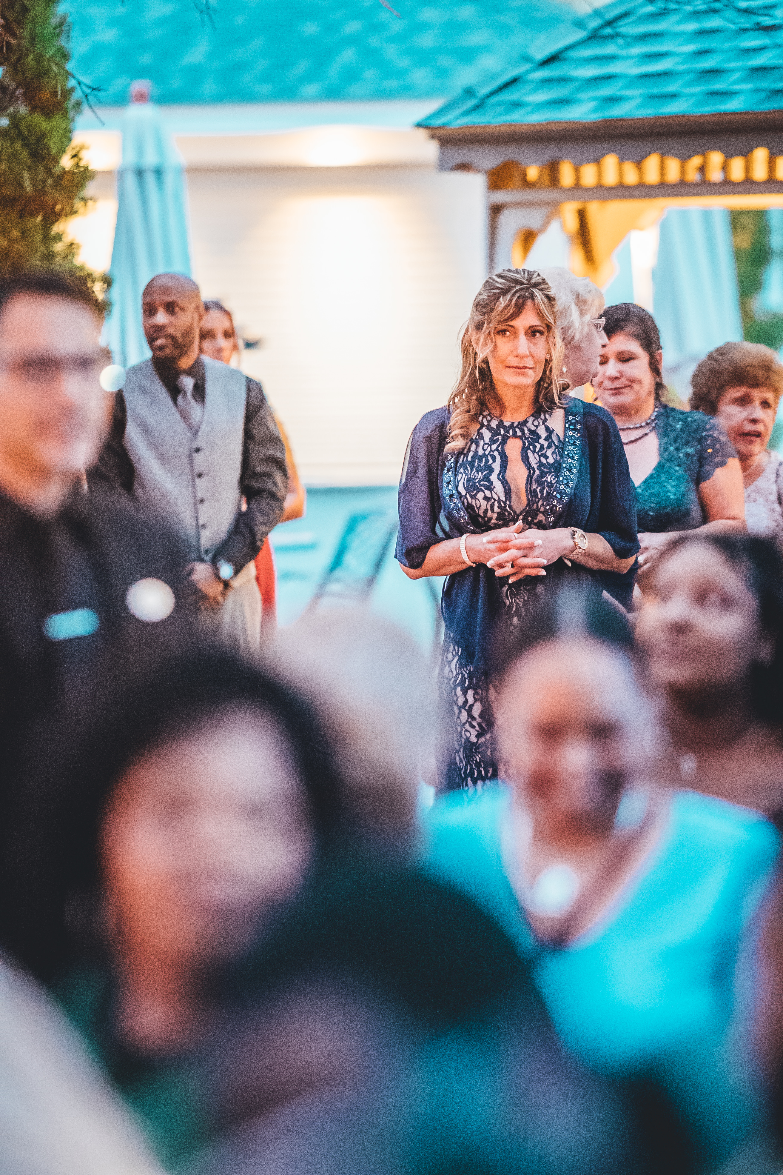 A woman in a navy lace dress stands among wedding guests at an outdoor poolside venue, speaking or being acknowledged during a reception moment. Guests in forma