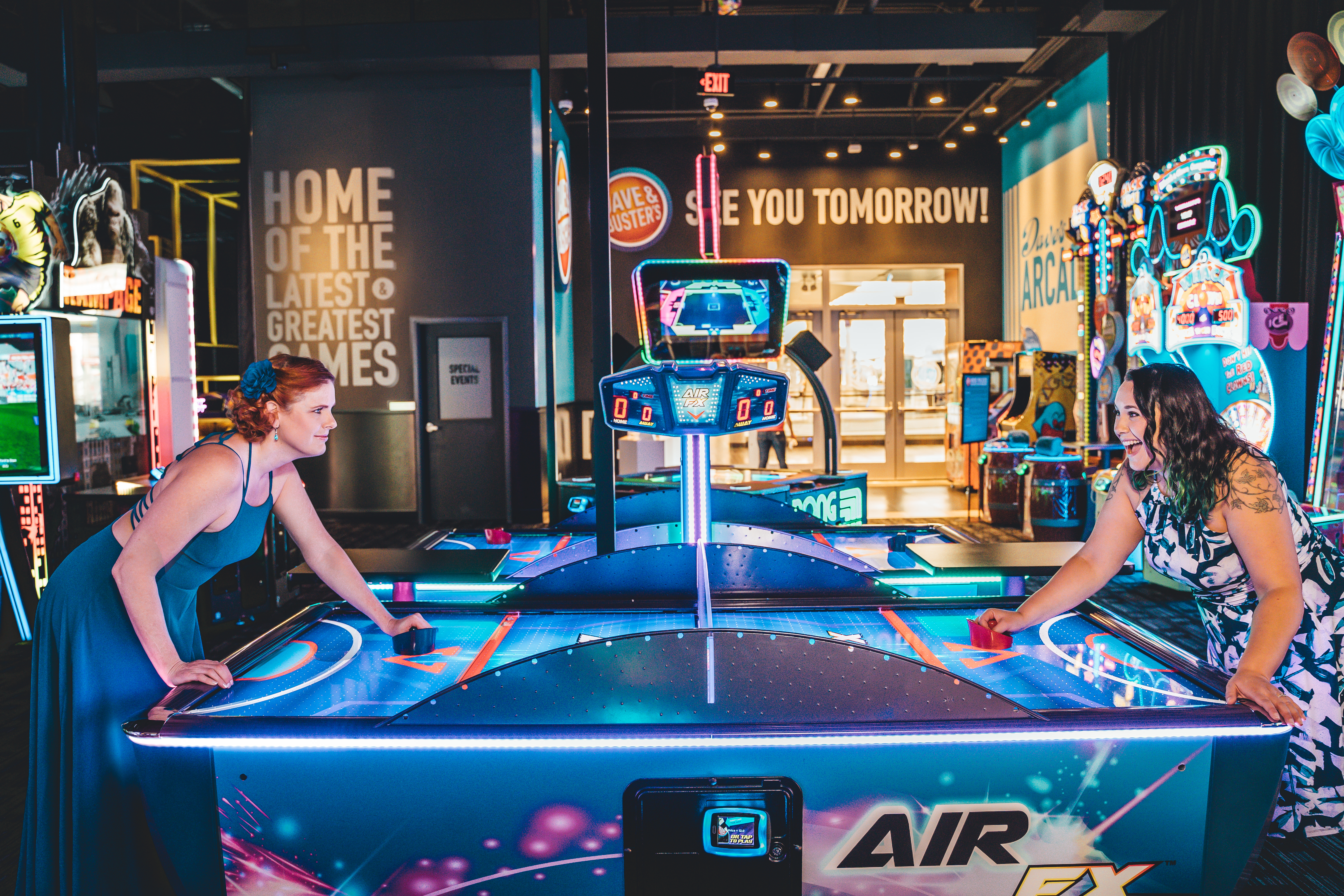 Two women play air hockey at a neon-lit arcade, facing each other across a glowing blue table. The arcade is filled with vintage gaming machines, neon signs reading 'HOME OF THE LATEST GREATEST GAMES' and 'SEE YOU TOMORROW,' and colorful game cabinets visible in the background. The setting is vibrant and playful, emphasizing their fun-loving personalities.