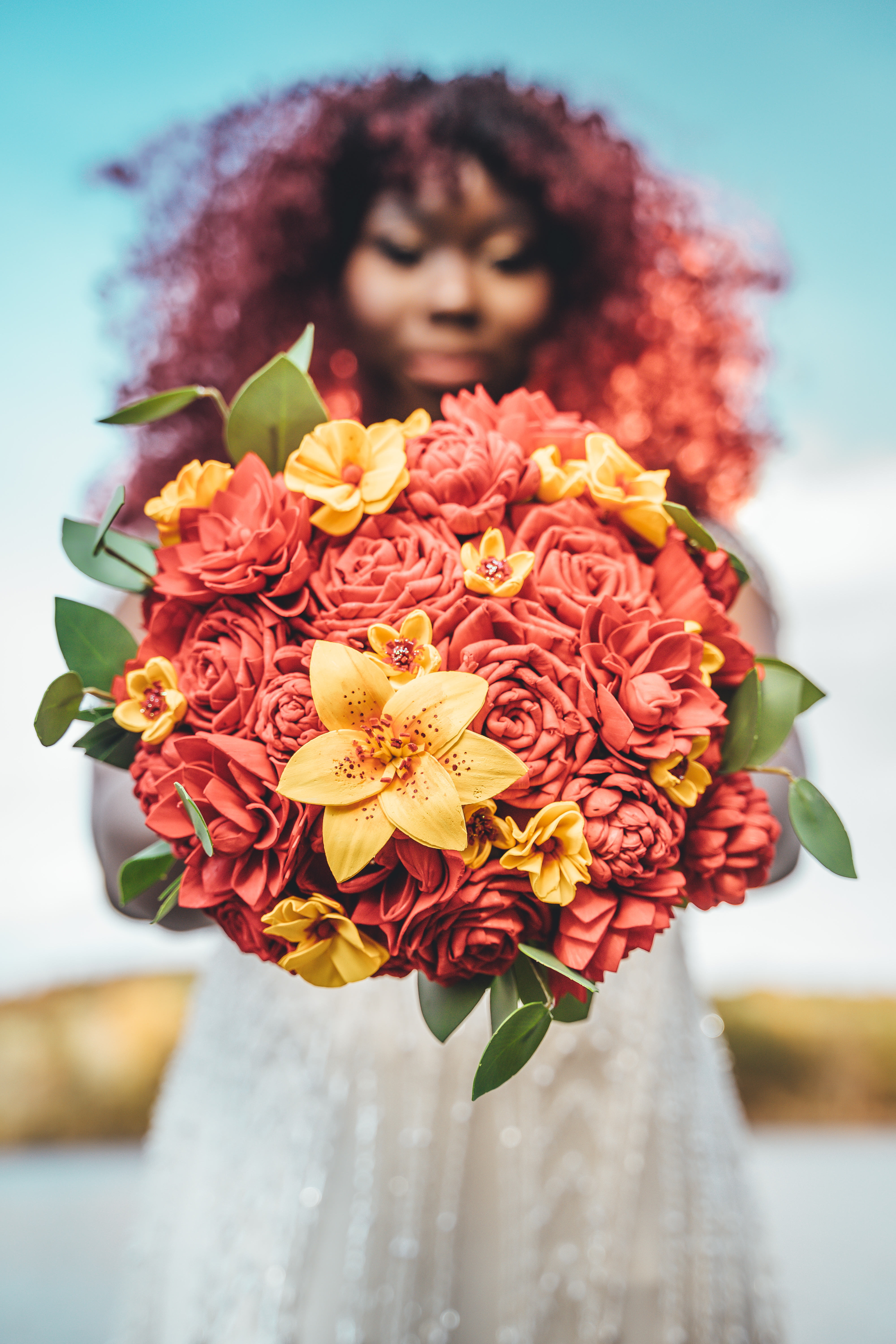 Bride holds a vibrant bouquet of coral roses and yellow lilies with green foliage, her face obscured behind the flowers.