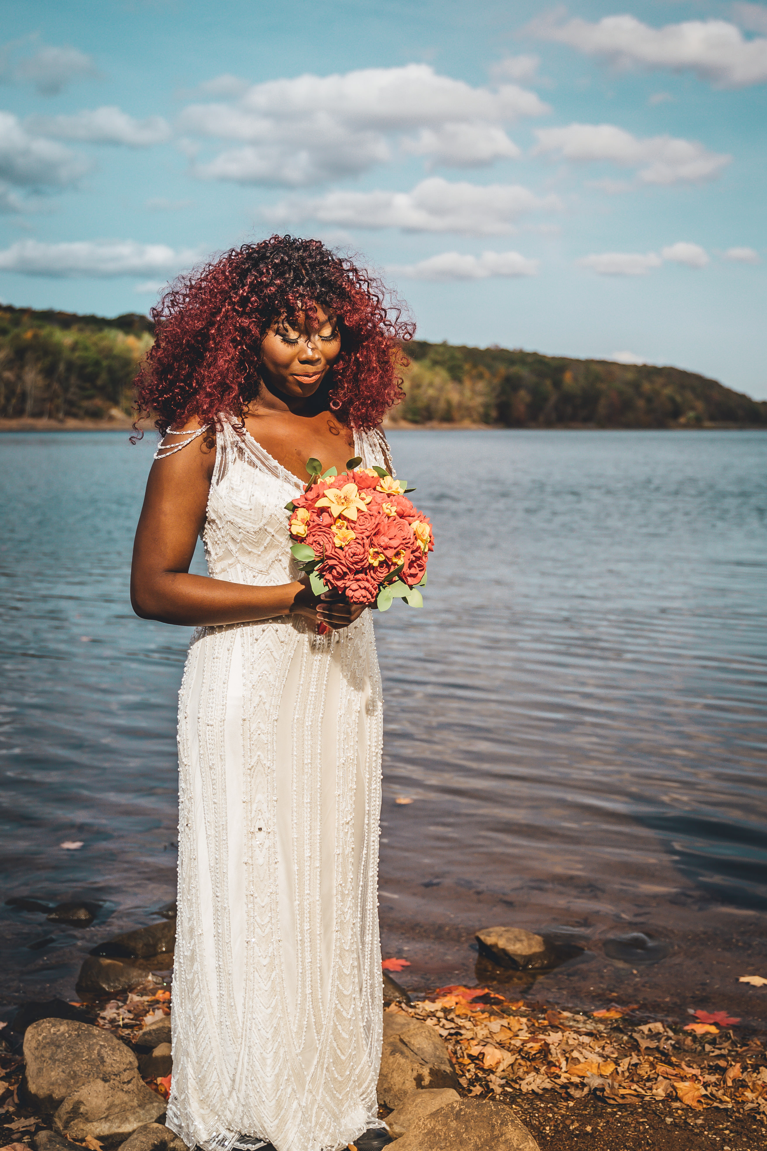 Bride stands alone at water's edge holding a vibrant coral and yellow bouquet, wearing a flowing white textured gown wit