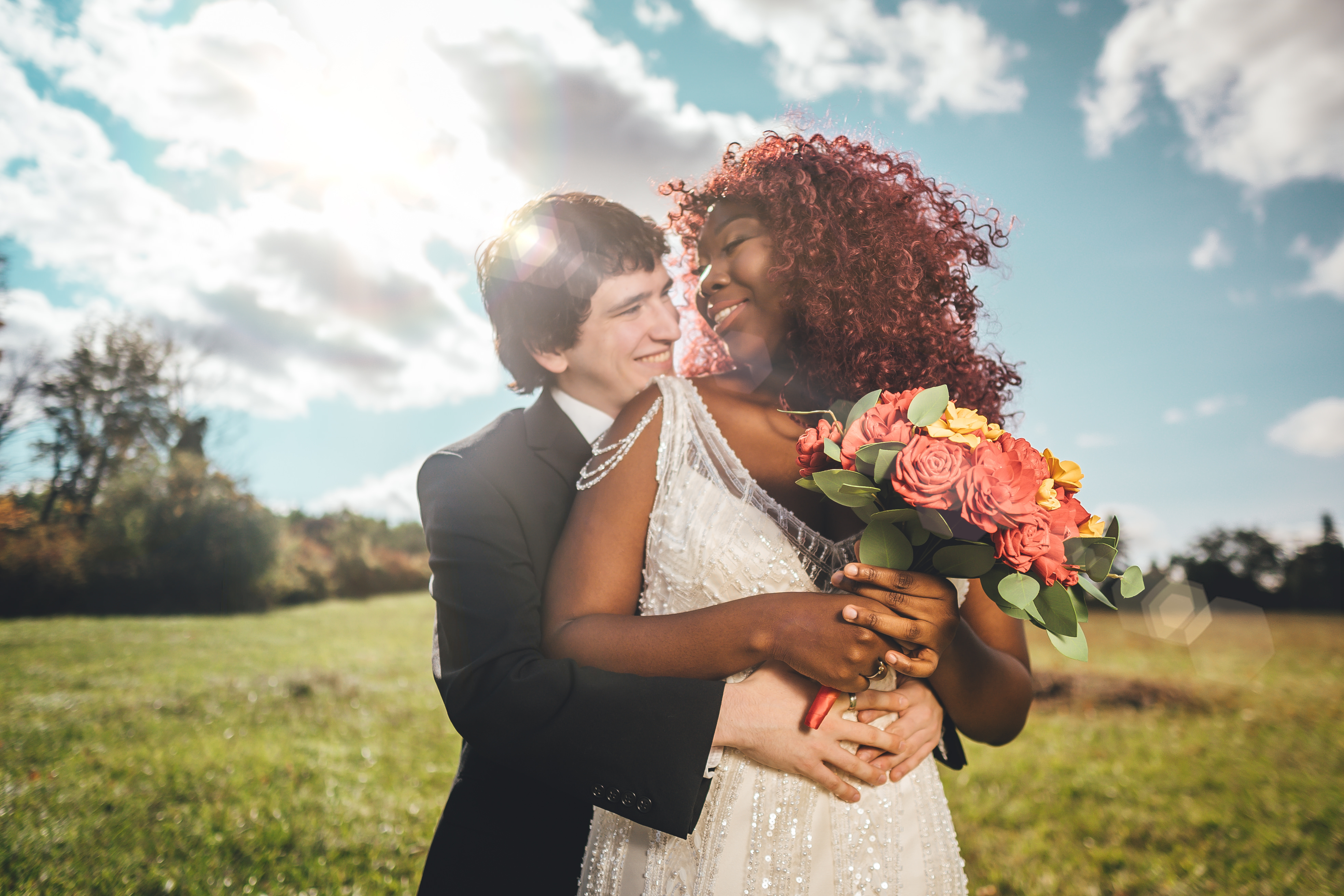 The couple embraces in an open field during golden hour, with the bride holding a coral and yellow rose bouquet. She wea