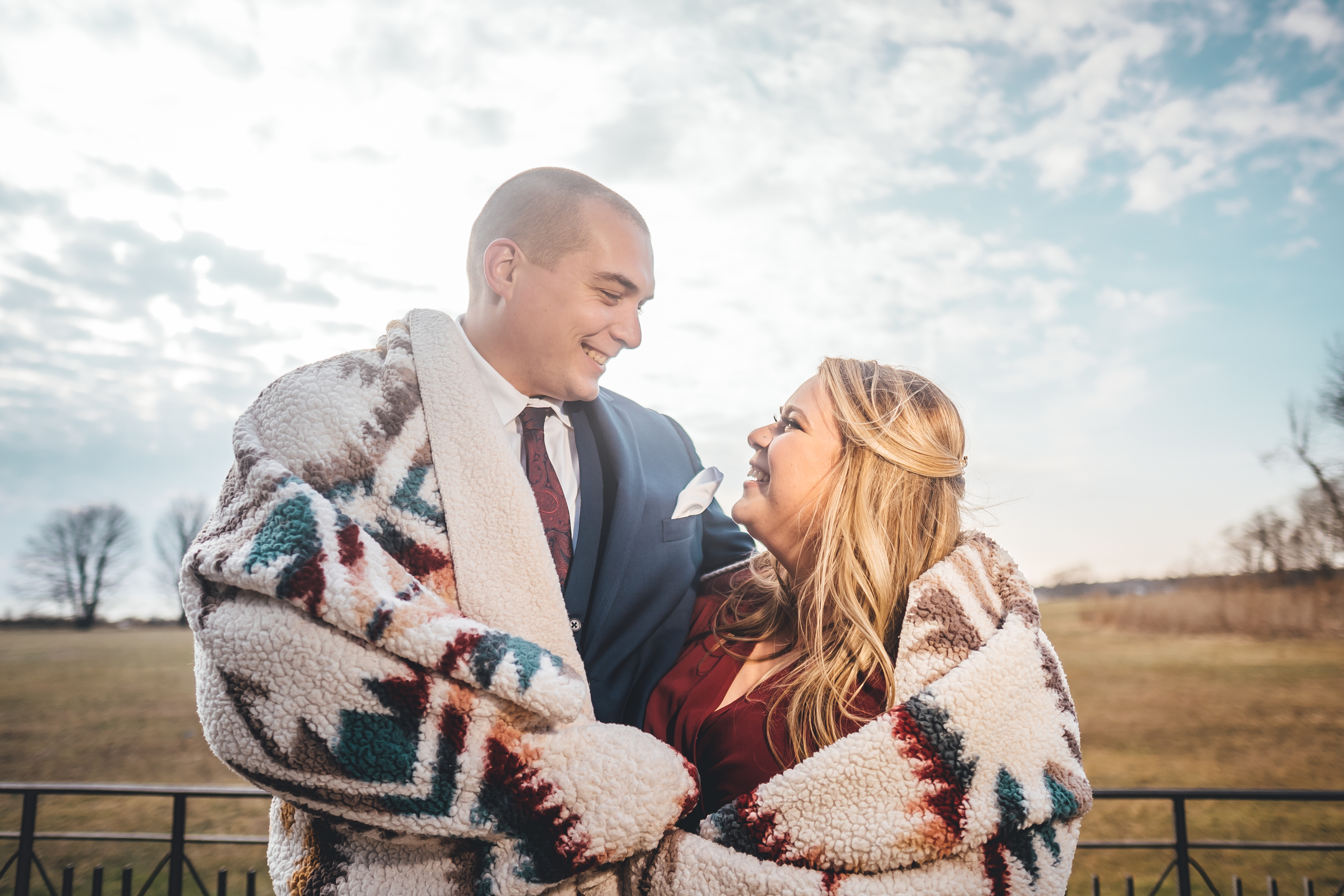 The couple stands close together outdoors wrapped in a cream-colored Pendleton-style blanket with geometric patterns in 