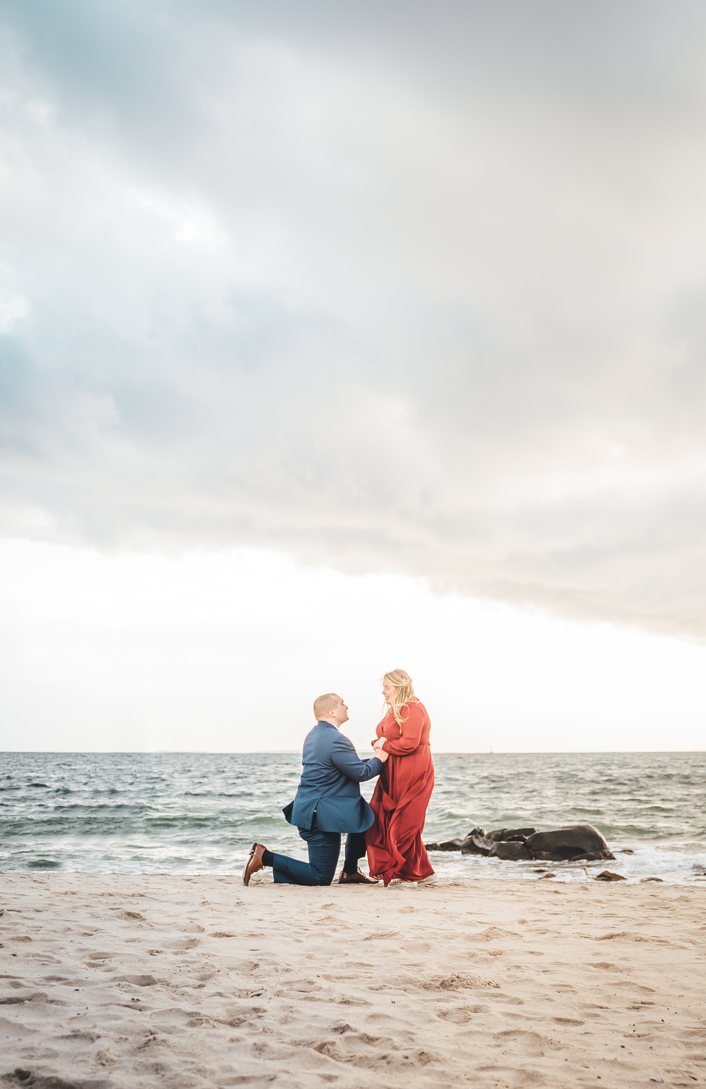 Groom in navy suit kneels on sand proposing to bride in flowing red/coral gown on a beach at Connecticut shoreline. Over