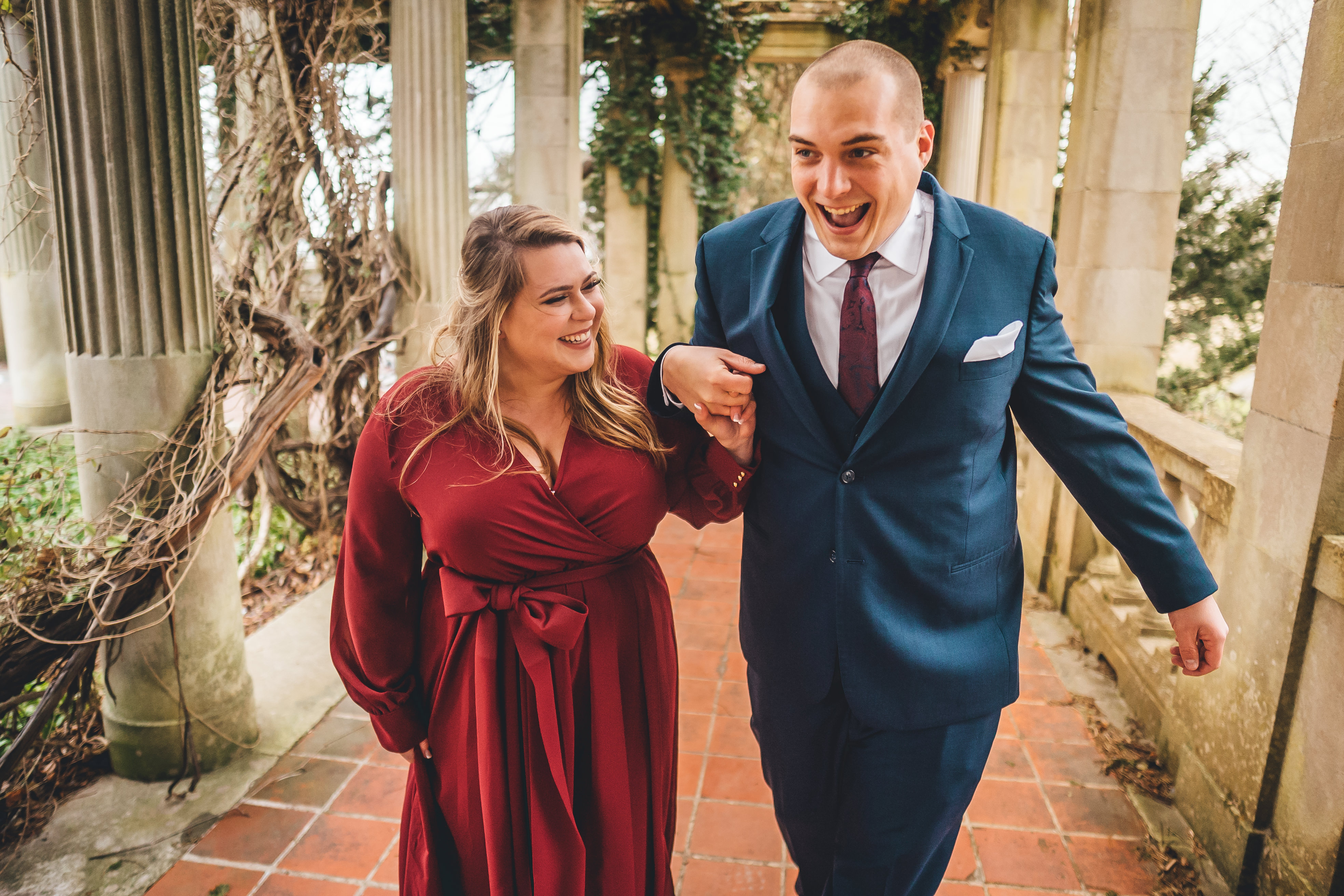 The groom in a navy blue suit and burgundy tie walks arm-in-arm with the bride down a classical colonnade. The bride wea