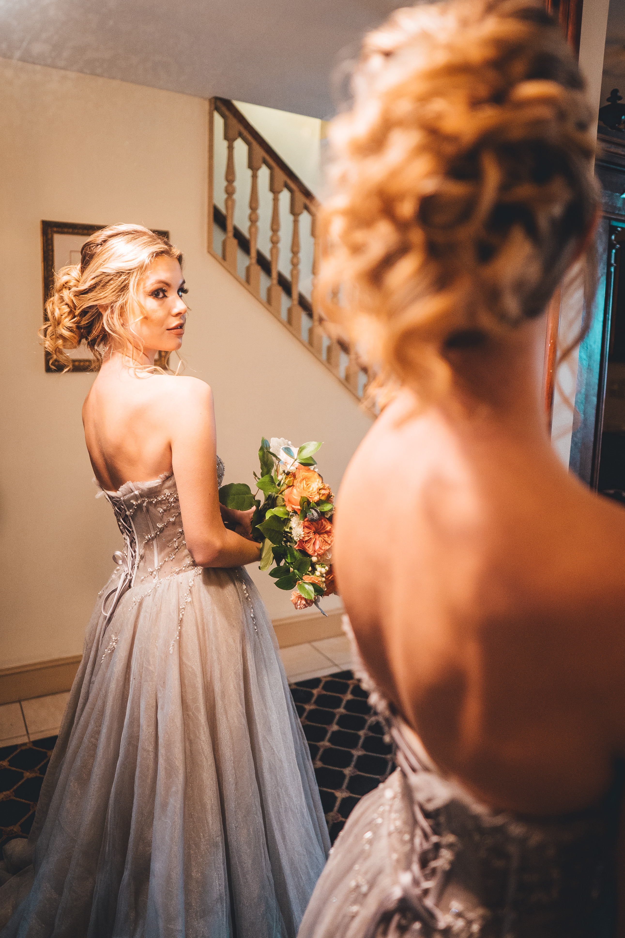 Bride in a gray strapless gown with beaded bodice and tulle skirt holds a peach and coral bouquet while gazing over her shoulder at a mirror reflection. She has