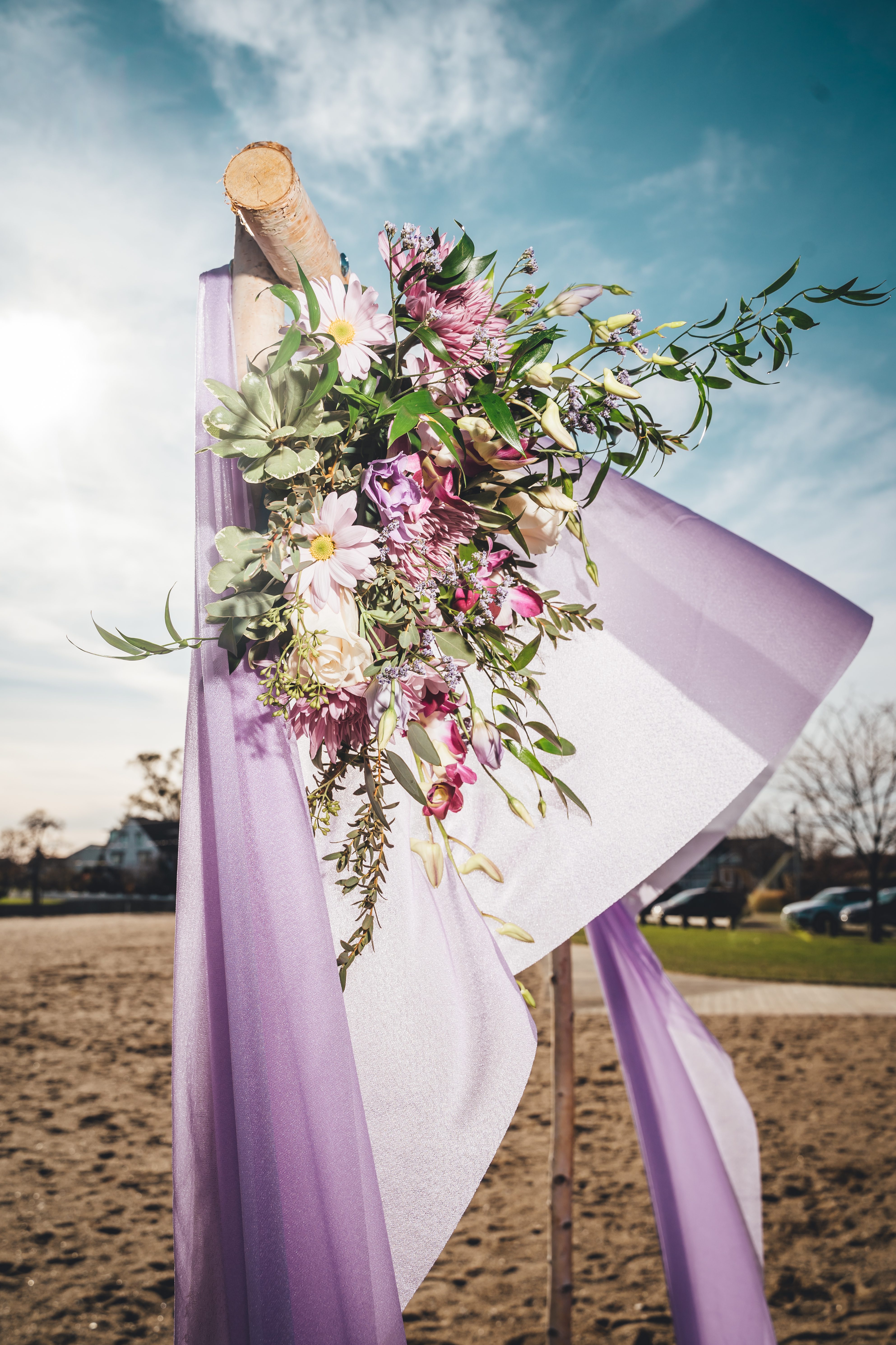 A decorative ceremony arch draped in lavender and white fabric is adorned with an asymmetrical floral arrangement featuring pink dahlias, white daisies, burgund