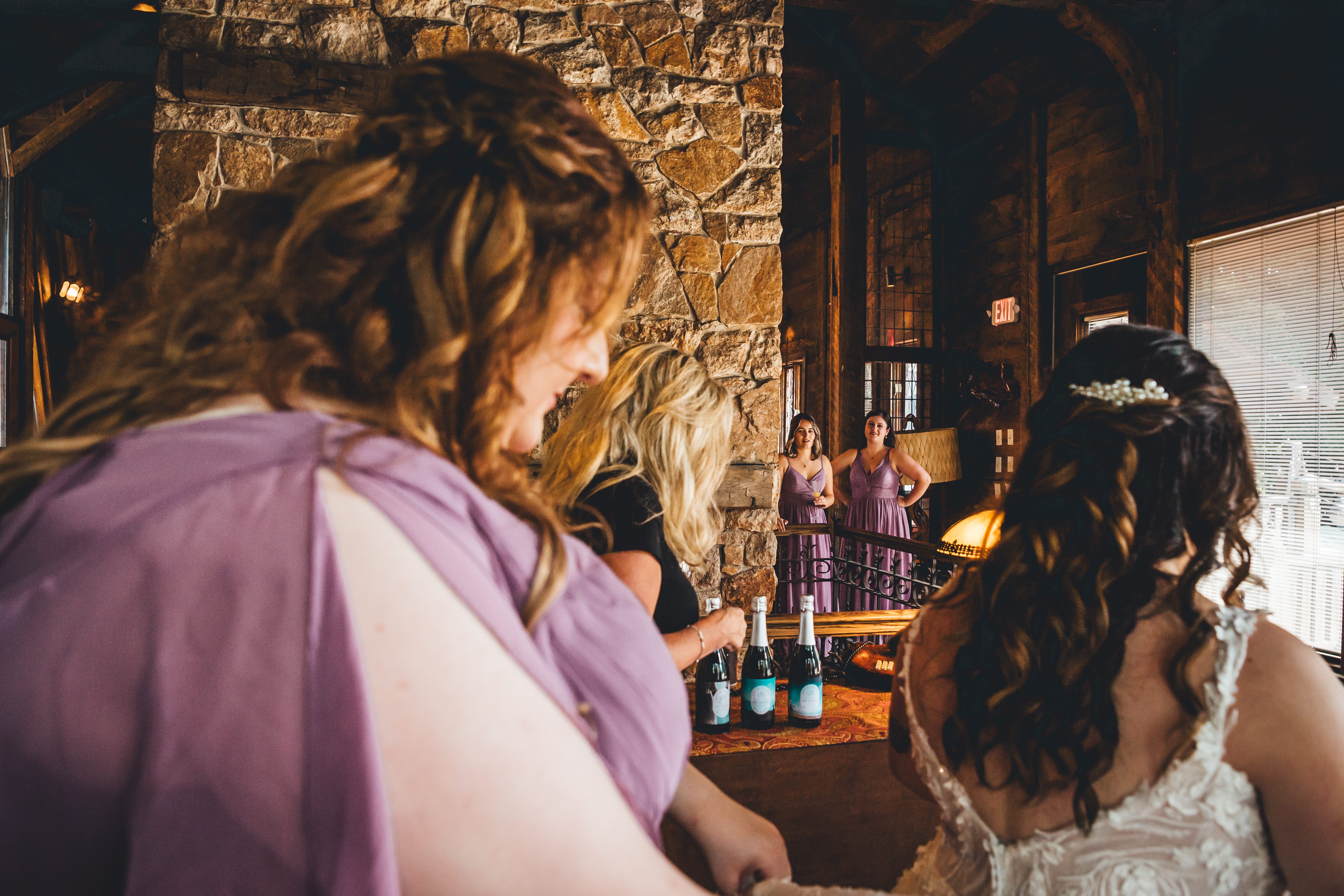 The bride in a white lace gown mingles with bridesmaids in lavender dresses in an elegant stone interior venue. Beer bottles are visible on a table, suggesting 