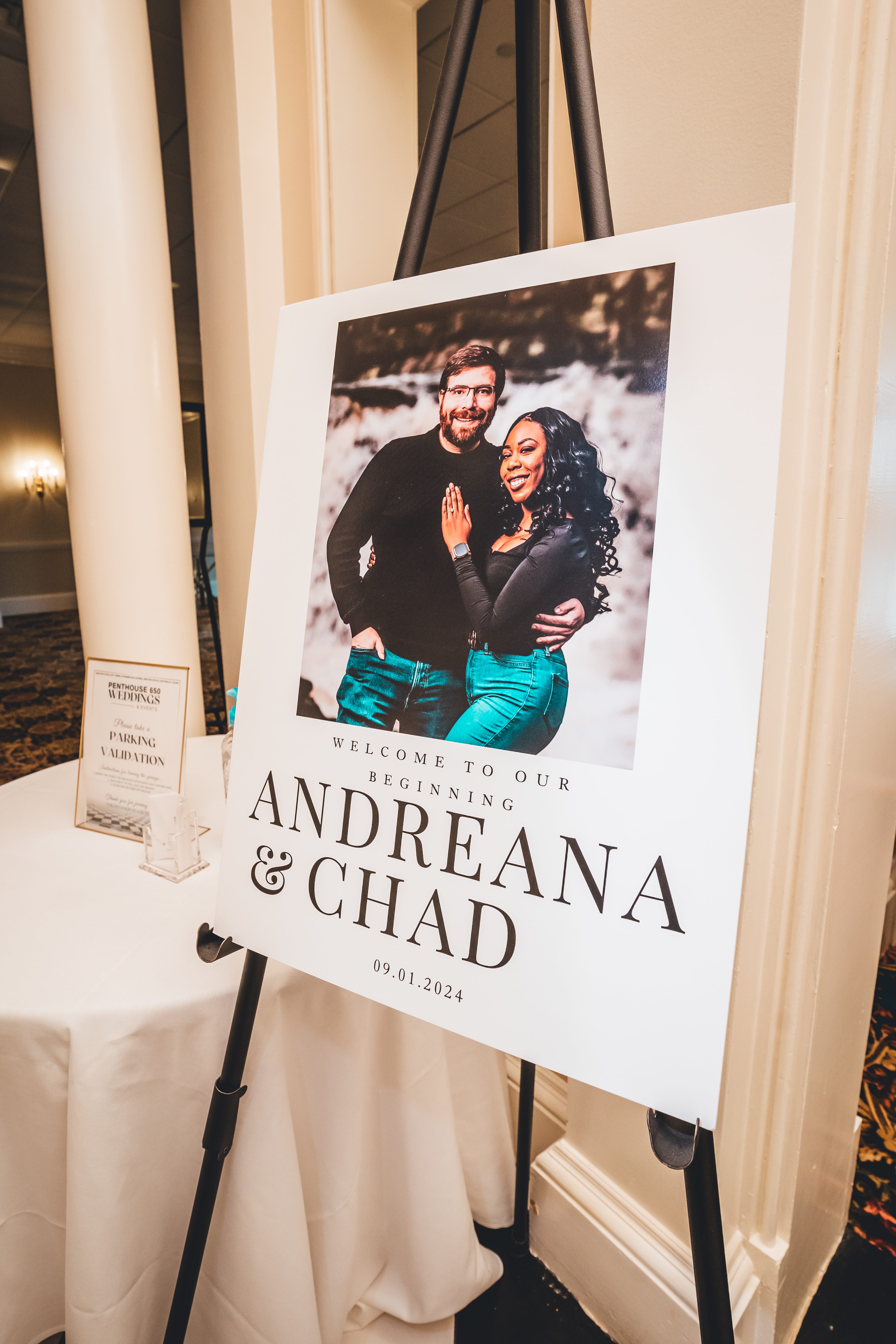 A large welcome sign featuring an engagement photo of the couple is displayed on an easel at the wedding reception entrance. The sign reads 'WELCOME TO OUR BEGI