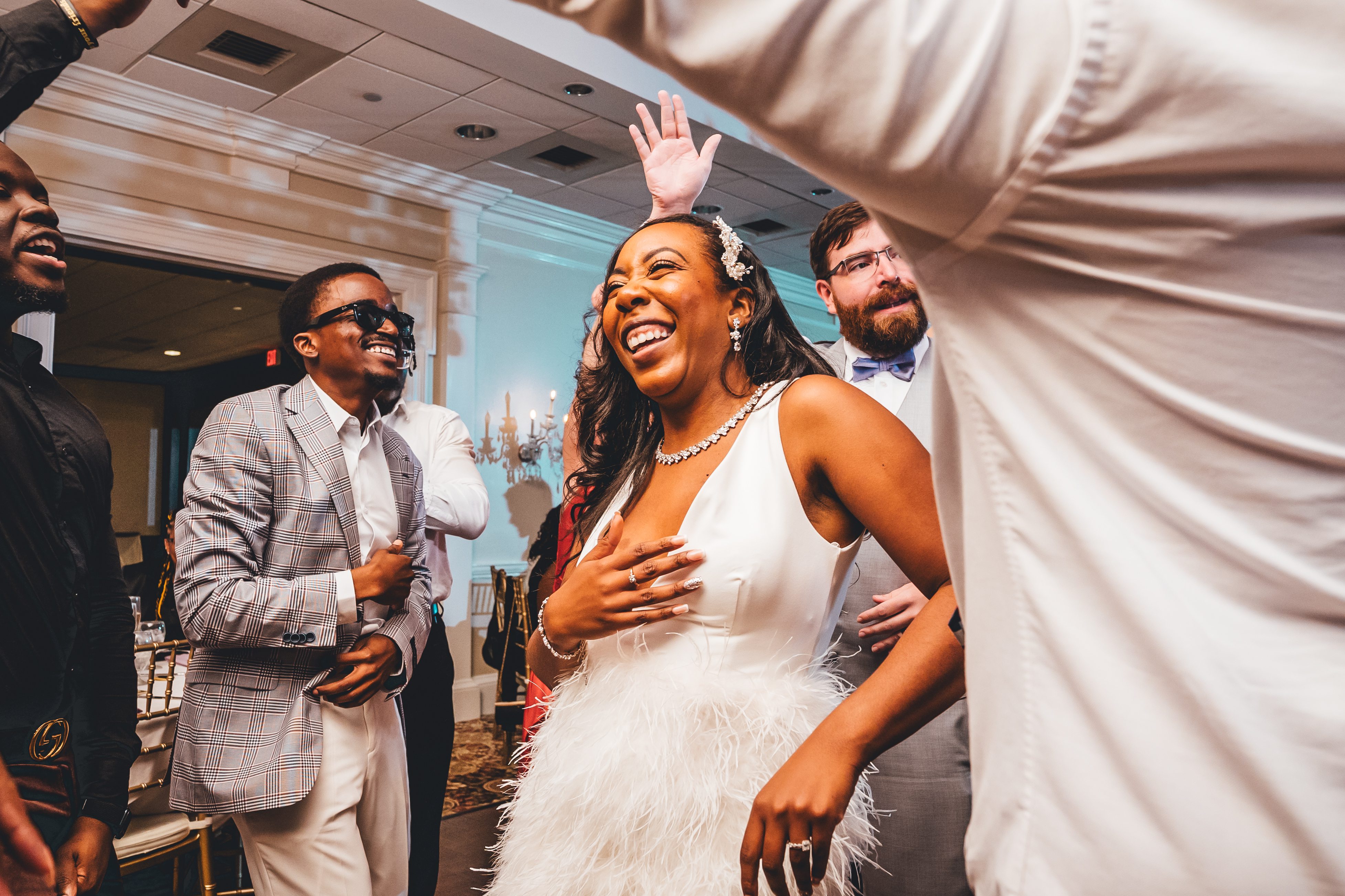 The bride in a white feathered gown with a flower crown celebrates joyfully with wedding guests indoors. A Disney castle backdrop is visible behind the group, i