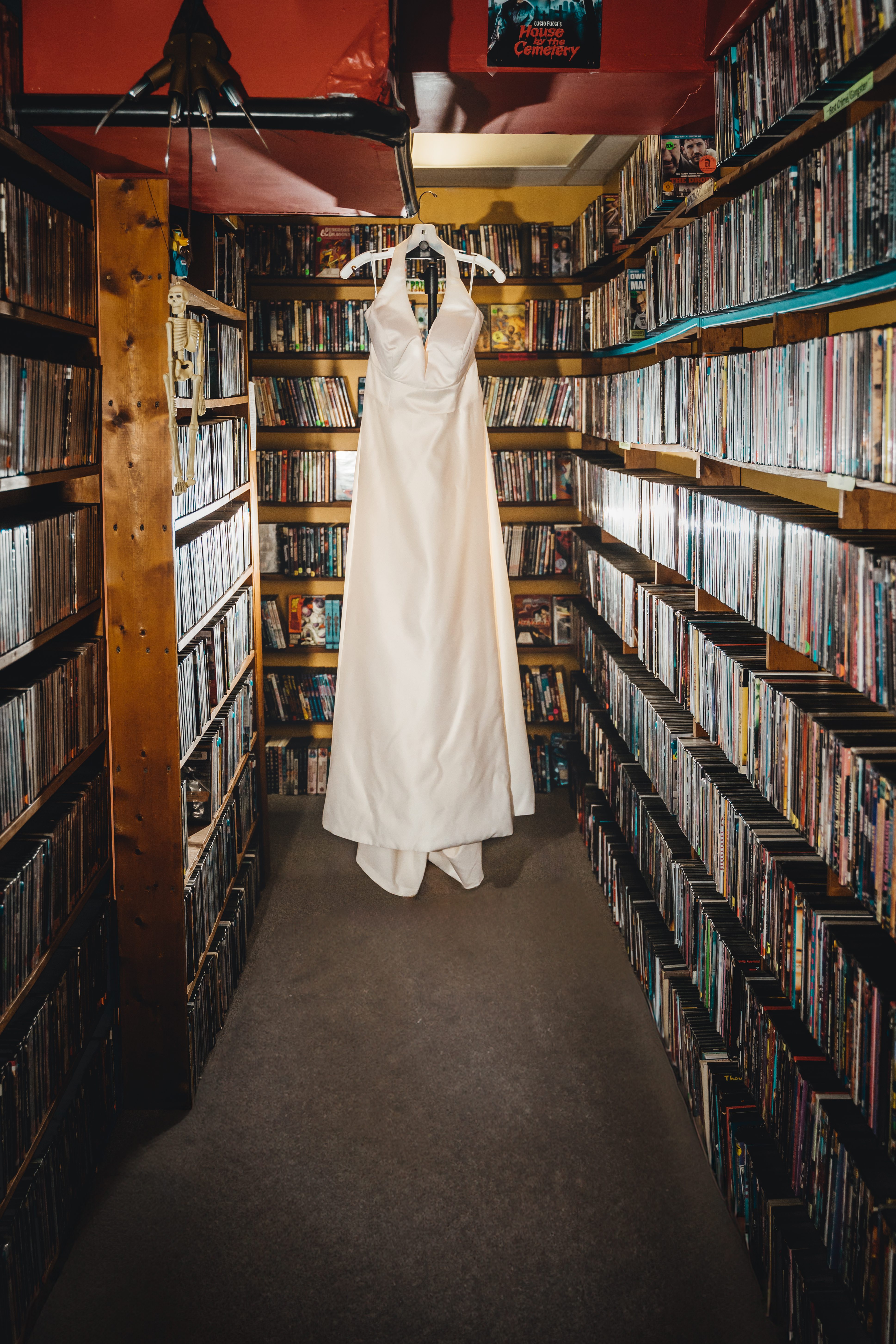 The bride's white wedding dress hangs on display between towering shelves of DVDs and Blu-rays in what appears to be a personal media collection room. The dress