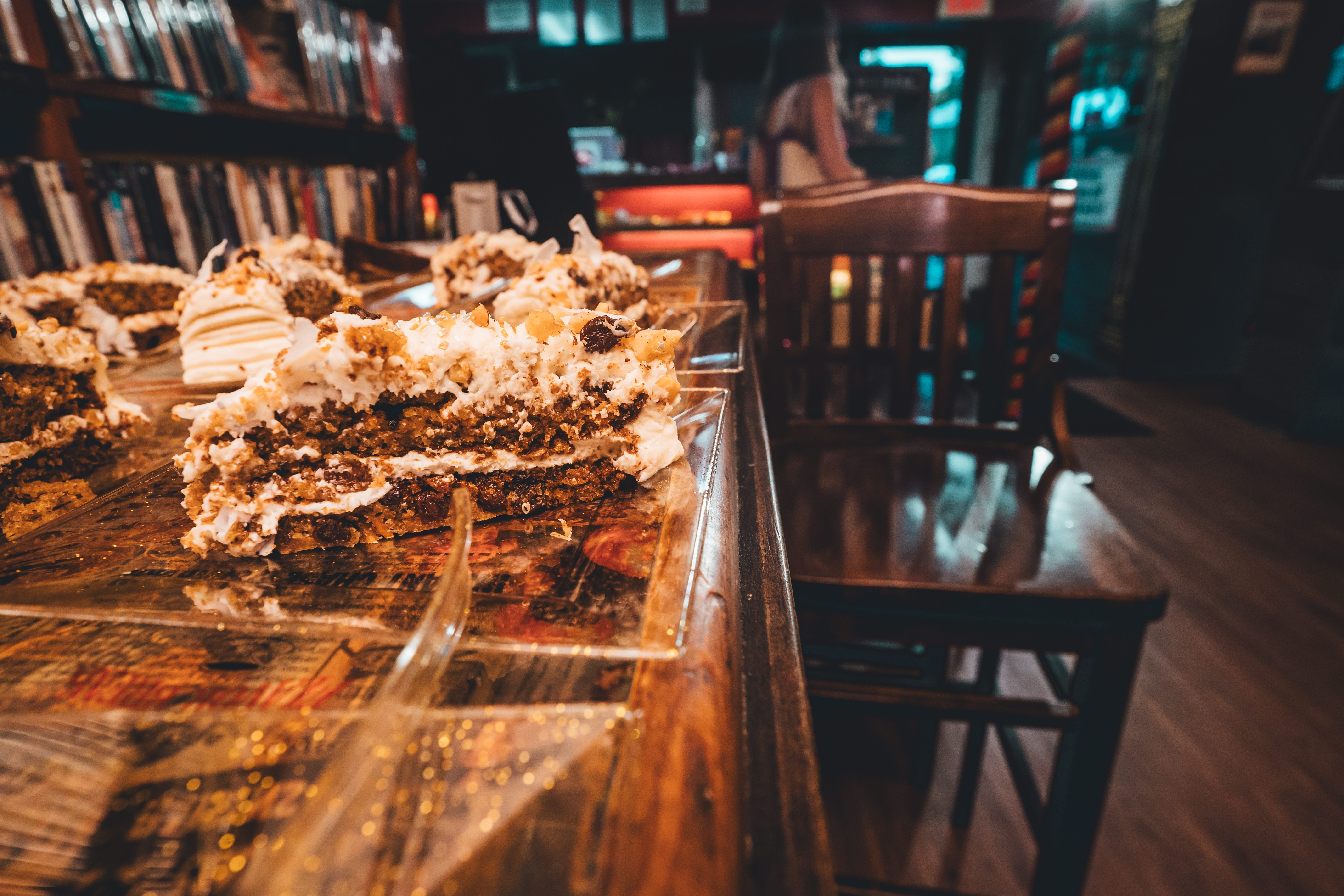Close-up detail shot of layered cake slices with cream frosting and crumb topping arranged on a wooden table. Bookshelves filled with volumes line the backgroun