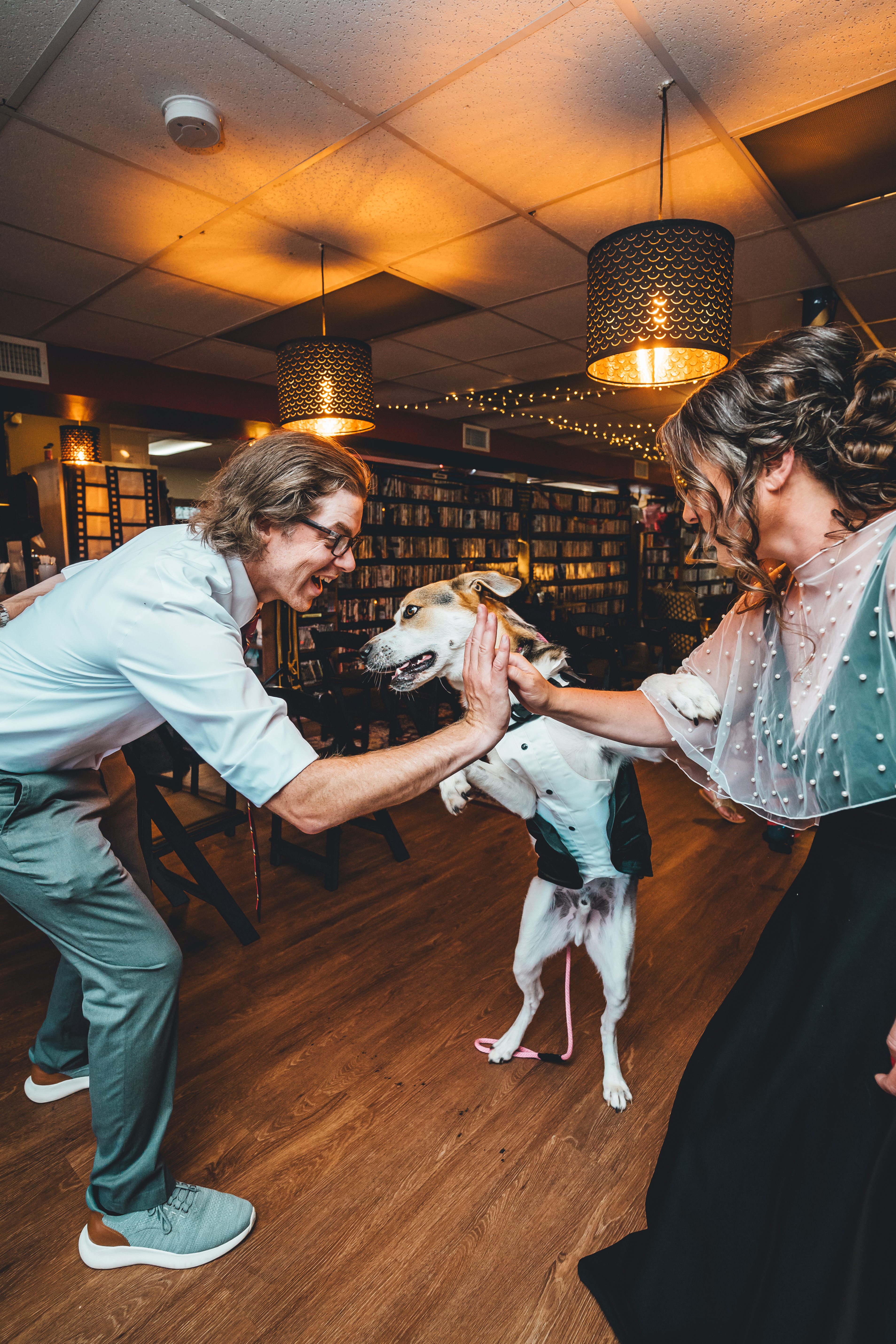 The groom and bride interact playfully with their dog in a cozy library venue filled with bookshelves. Both are smiling and petting the small tan and white dog 