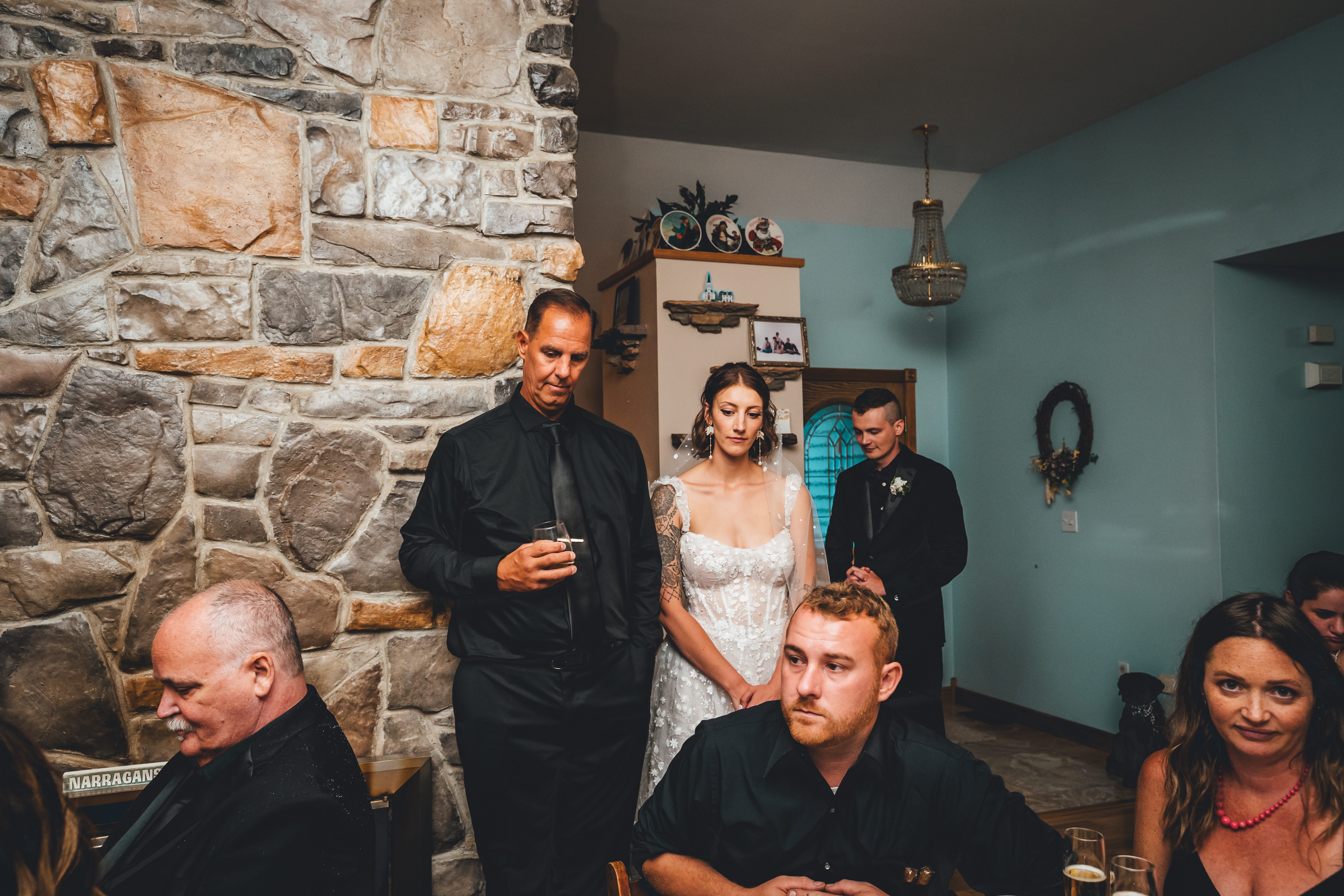 A bride in a beaded white gown stands with a groom in black and wedding guests in an eclectic indoor reception space. The venue features exposed stone walls, tu