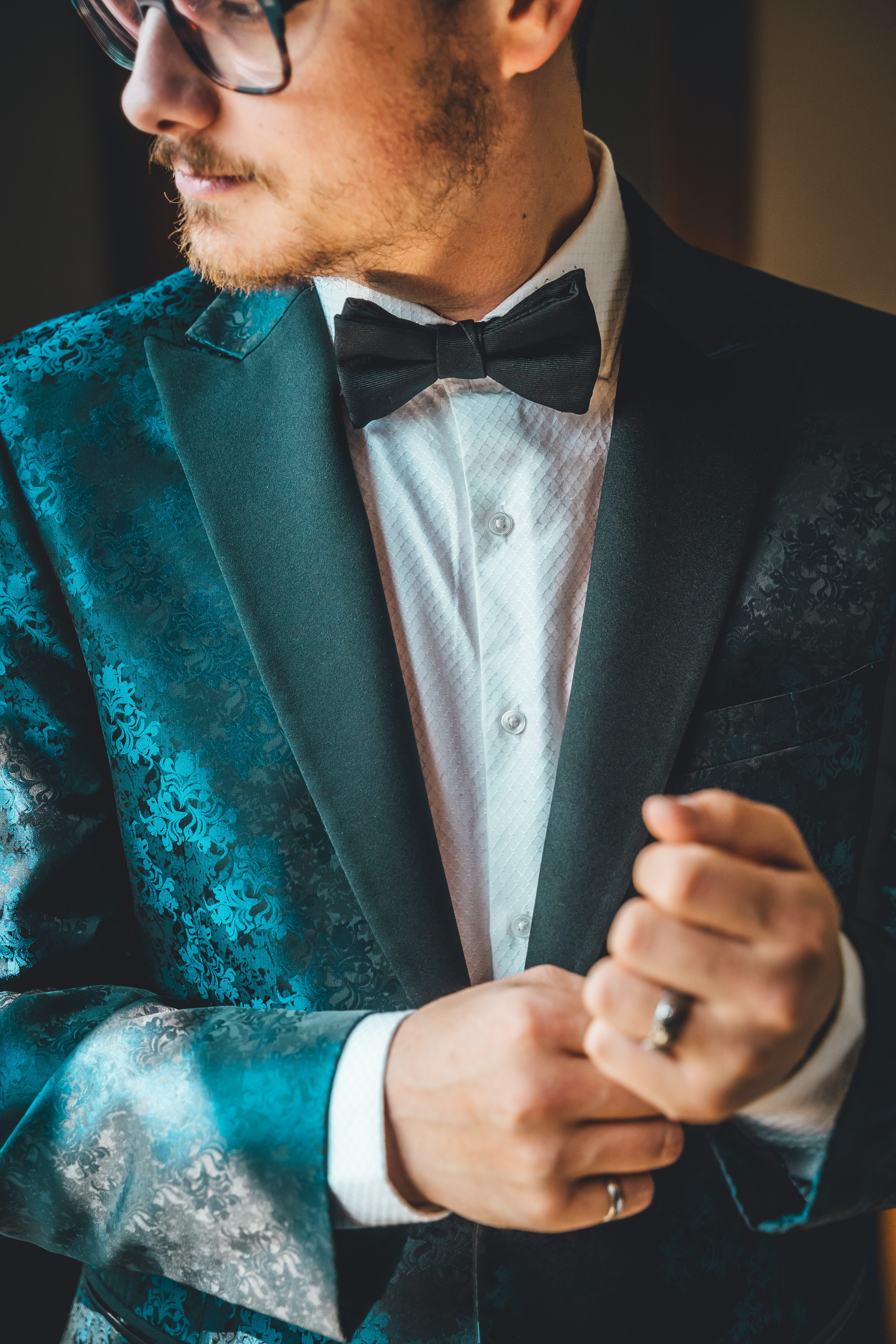 A groom in a teal floral jacquard tuxedo jacket adjusts his cuff
