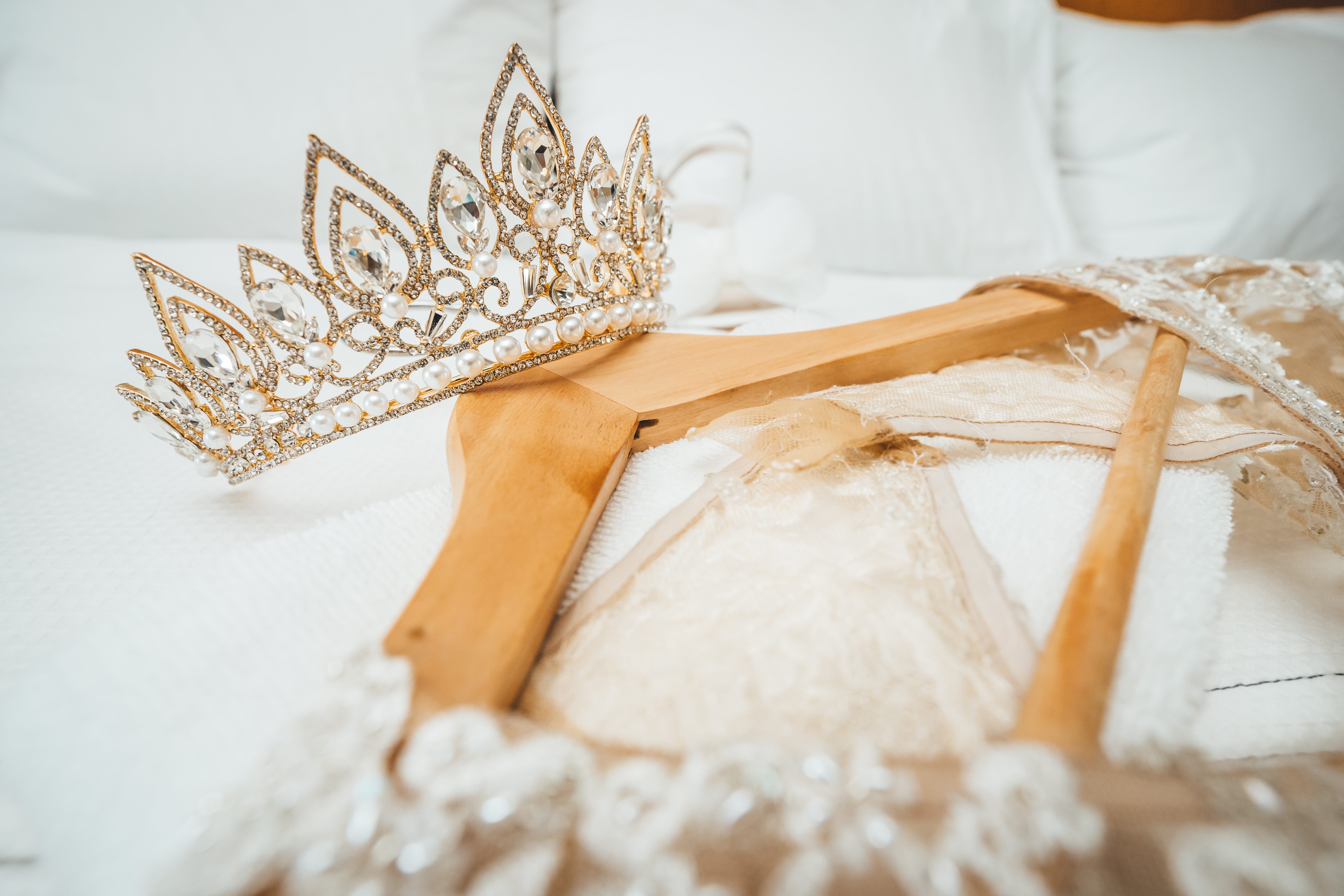 A crystal tiara resting on a lace wedding dress on its hanger