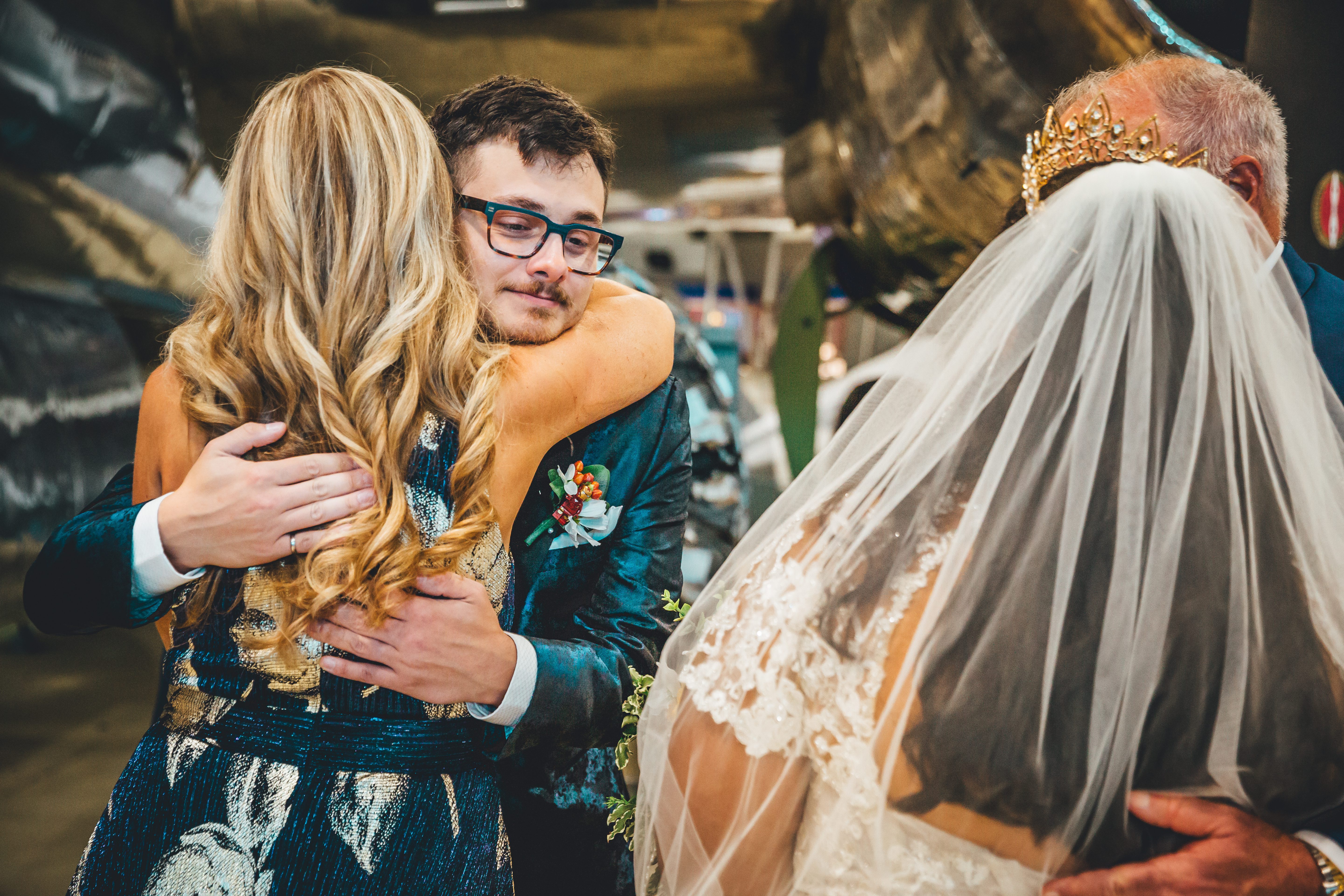The groom in an ornate teal and gold brocade jacket embraces the bride while greeting an older woman wearing a golden crown and ivory veil. The intimate moment 