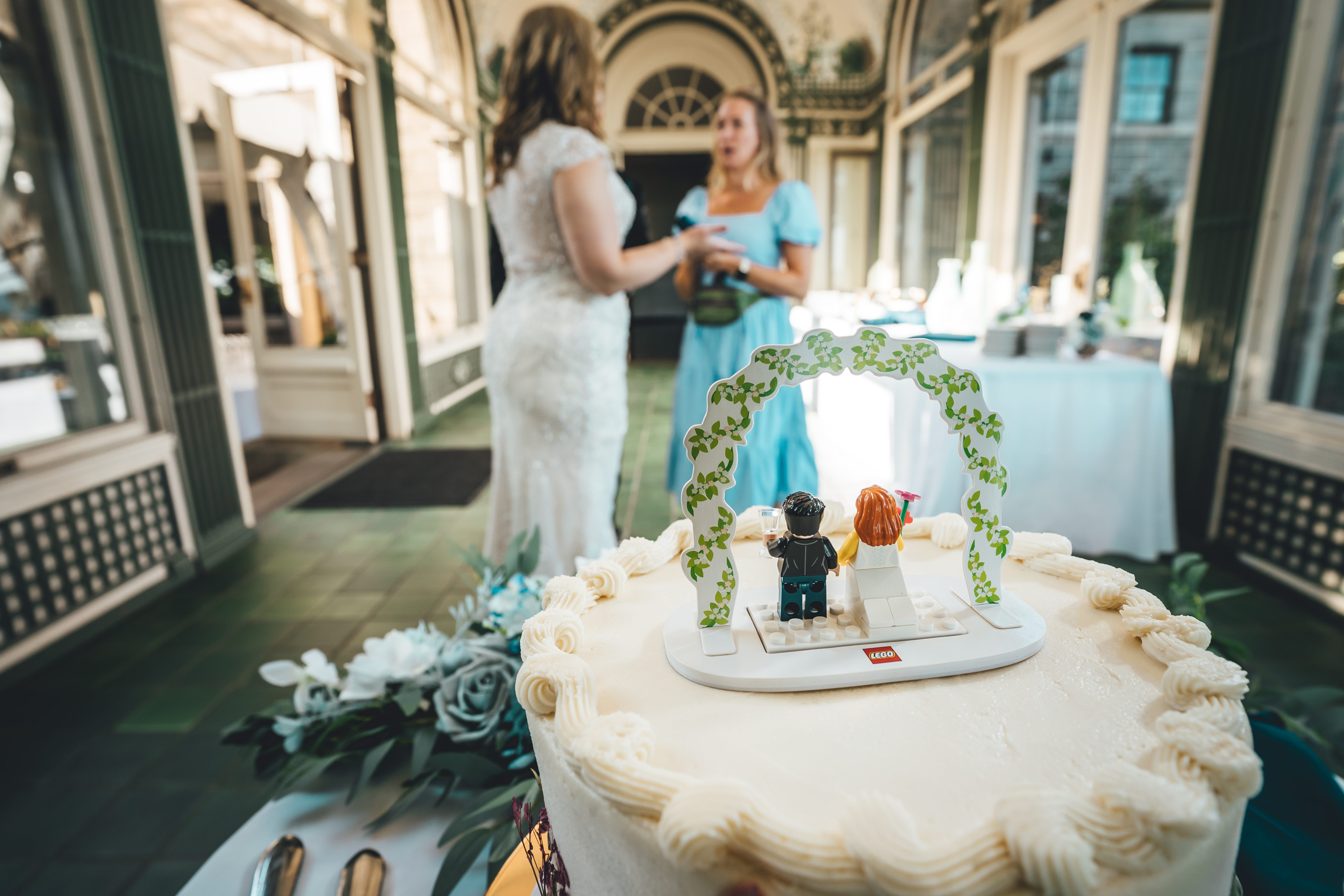 A wedding cake is prominently displayed with LEGO minifigure cake toppers of the bride and groom in formal attire, set under a decorative greenery arch. The bri