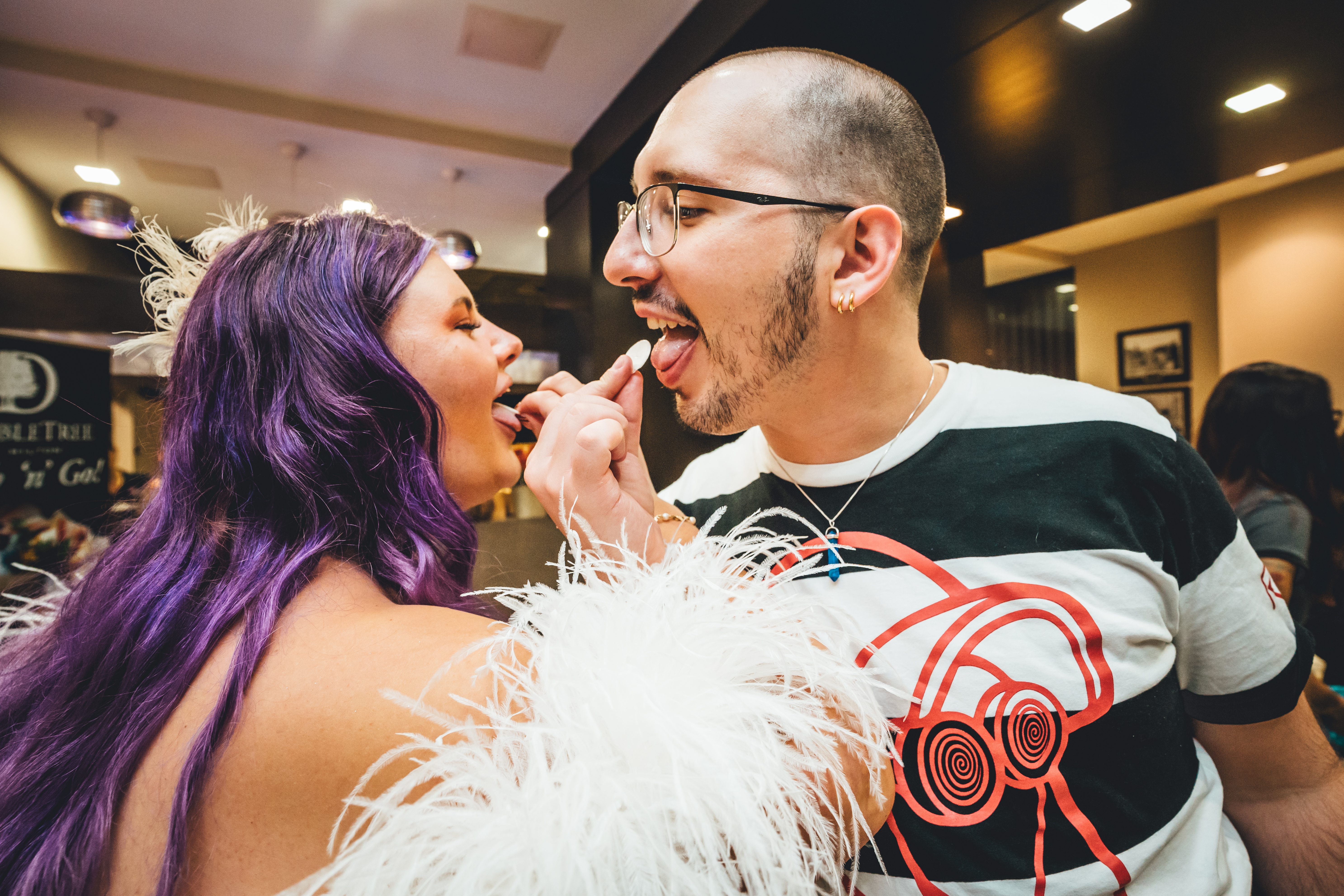 The groom feeds cake to the bride during the cake-cutting moment. The bride has vibrant purple hair and wears a white feathered dress; the groom wears a graphic