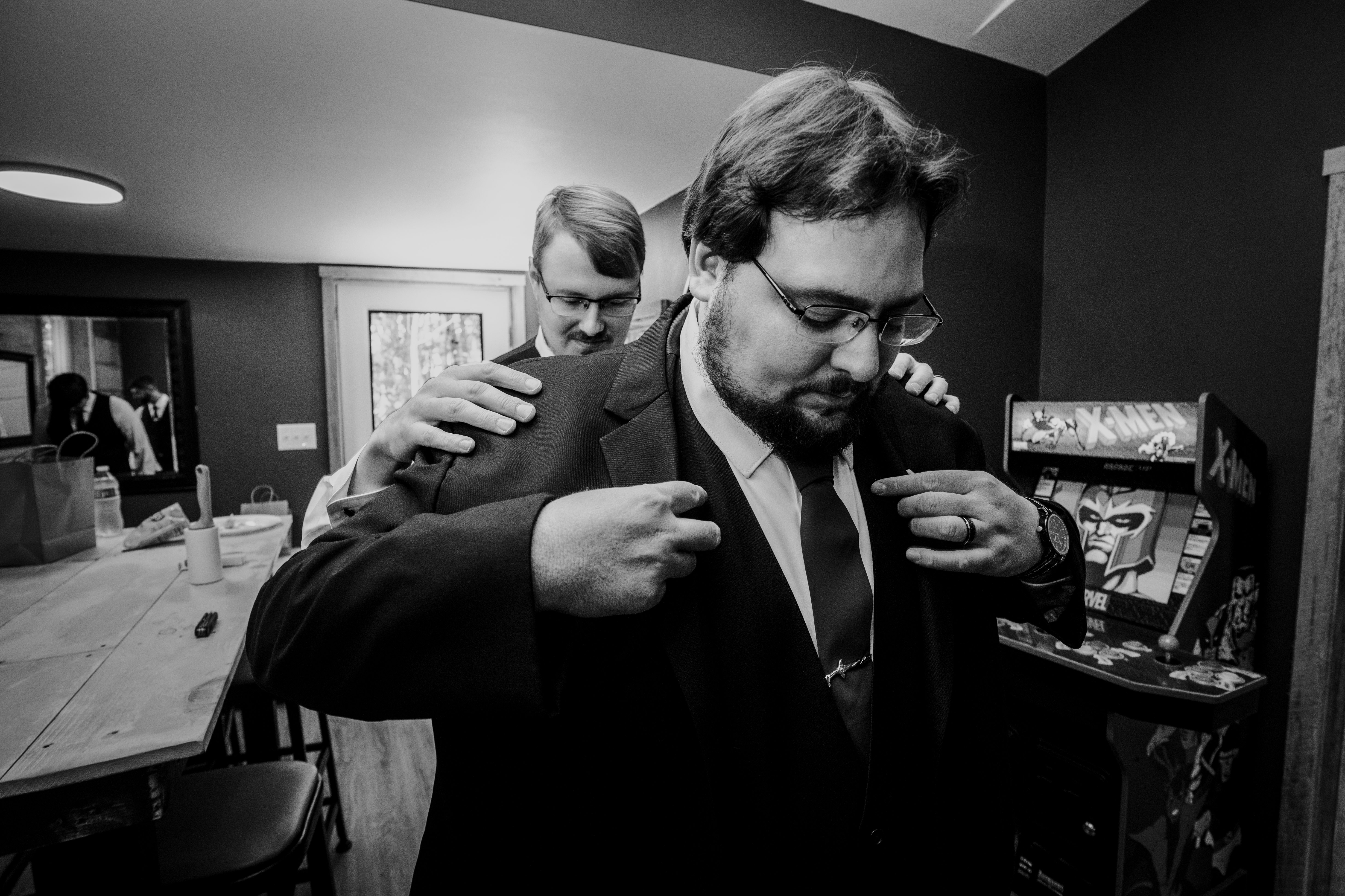 Two groomsmen in formal black suits examine comic book collectibles displayed on a shelf during getting-ready preparations. One man points at the comics while h