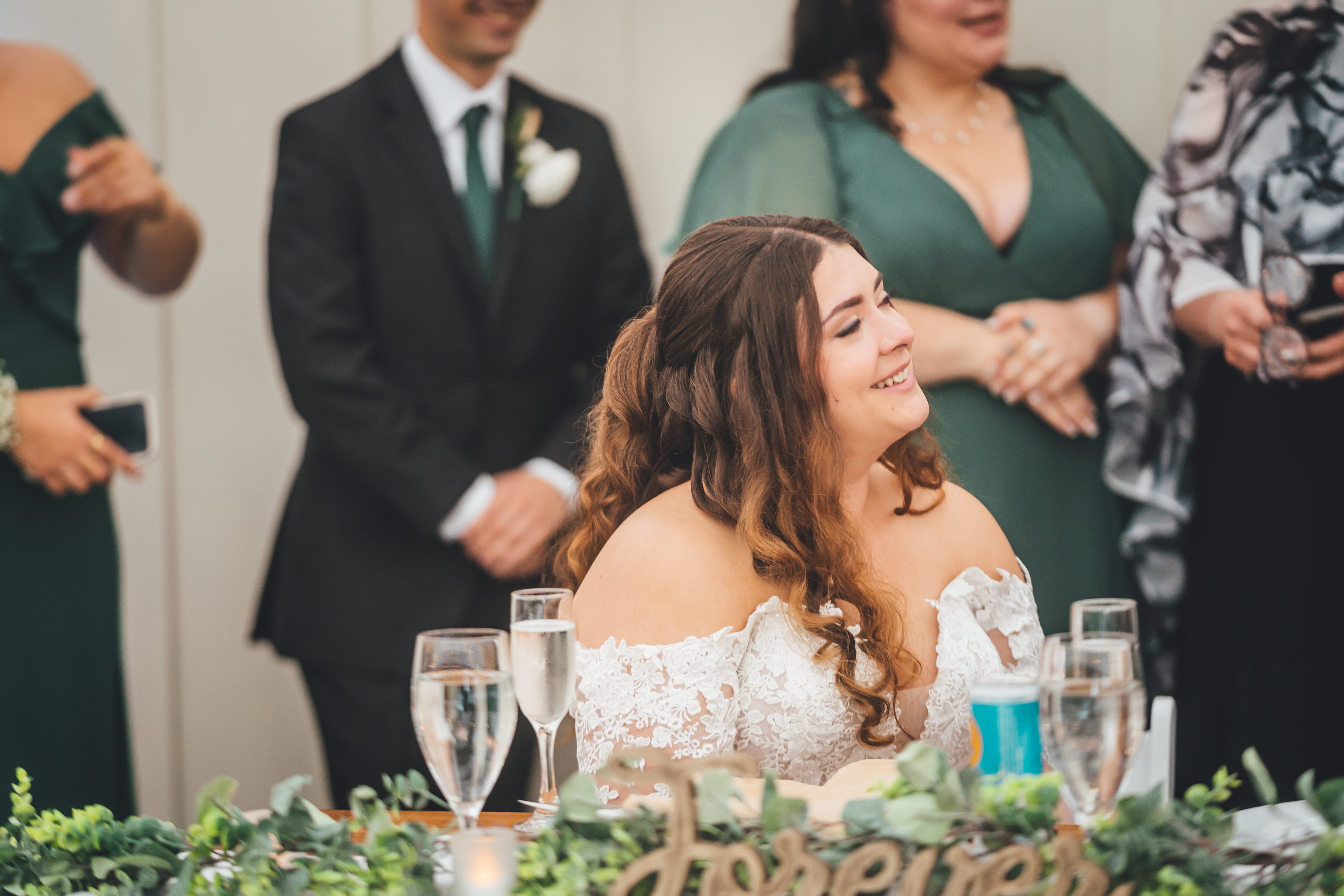 The bride sits at the reception table in her white lace off-shoulder gown, smiling joyfully while surrounded by wedding guests in formal attire. Behind her stan