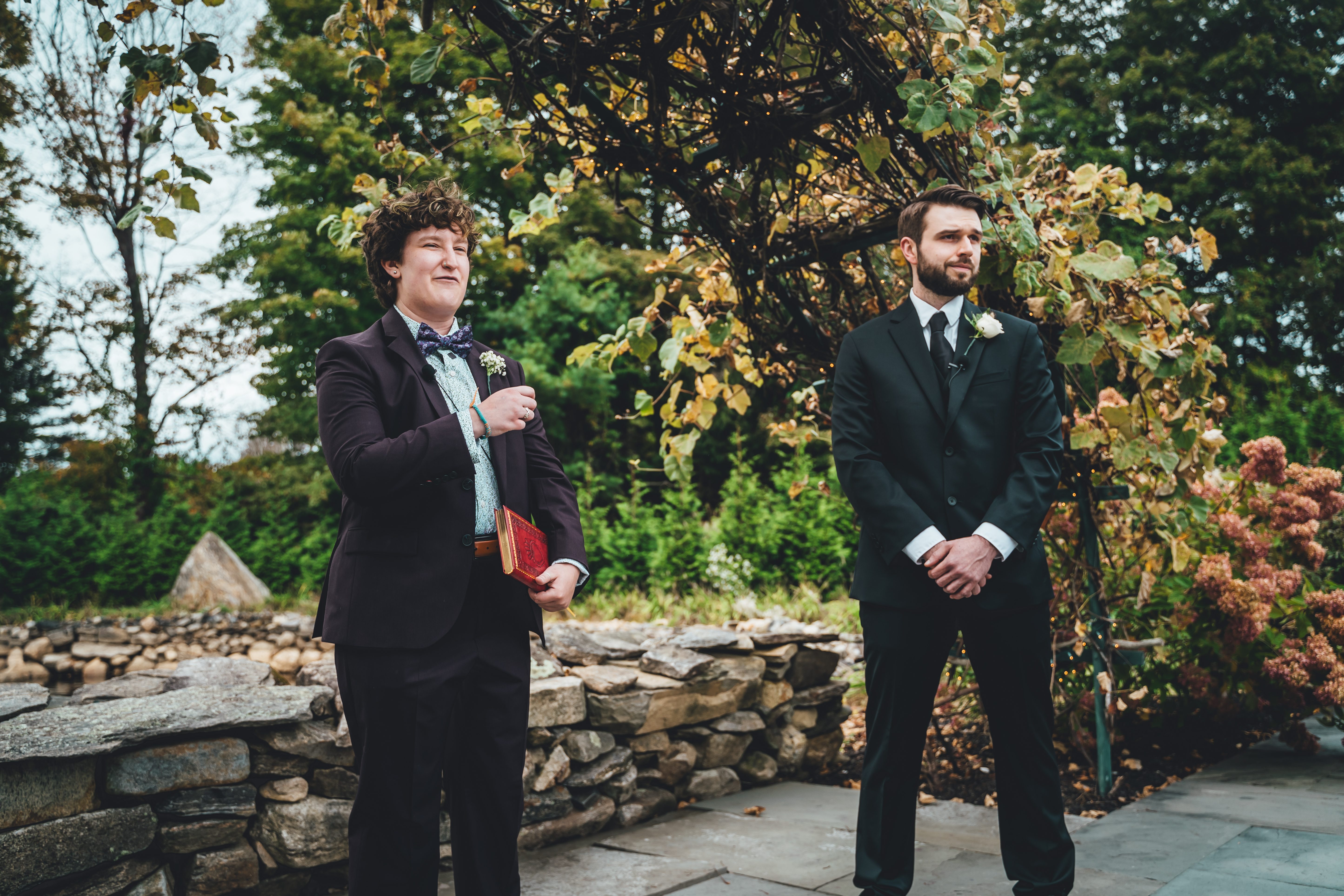 Two grooms stand in formal attire during an outdoor ceremony among stone walls and autumn foliage. The groom on the left