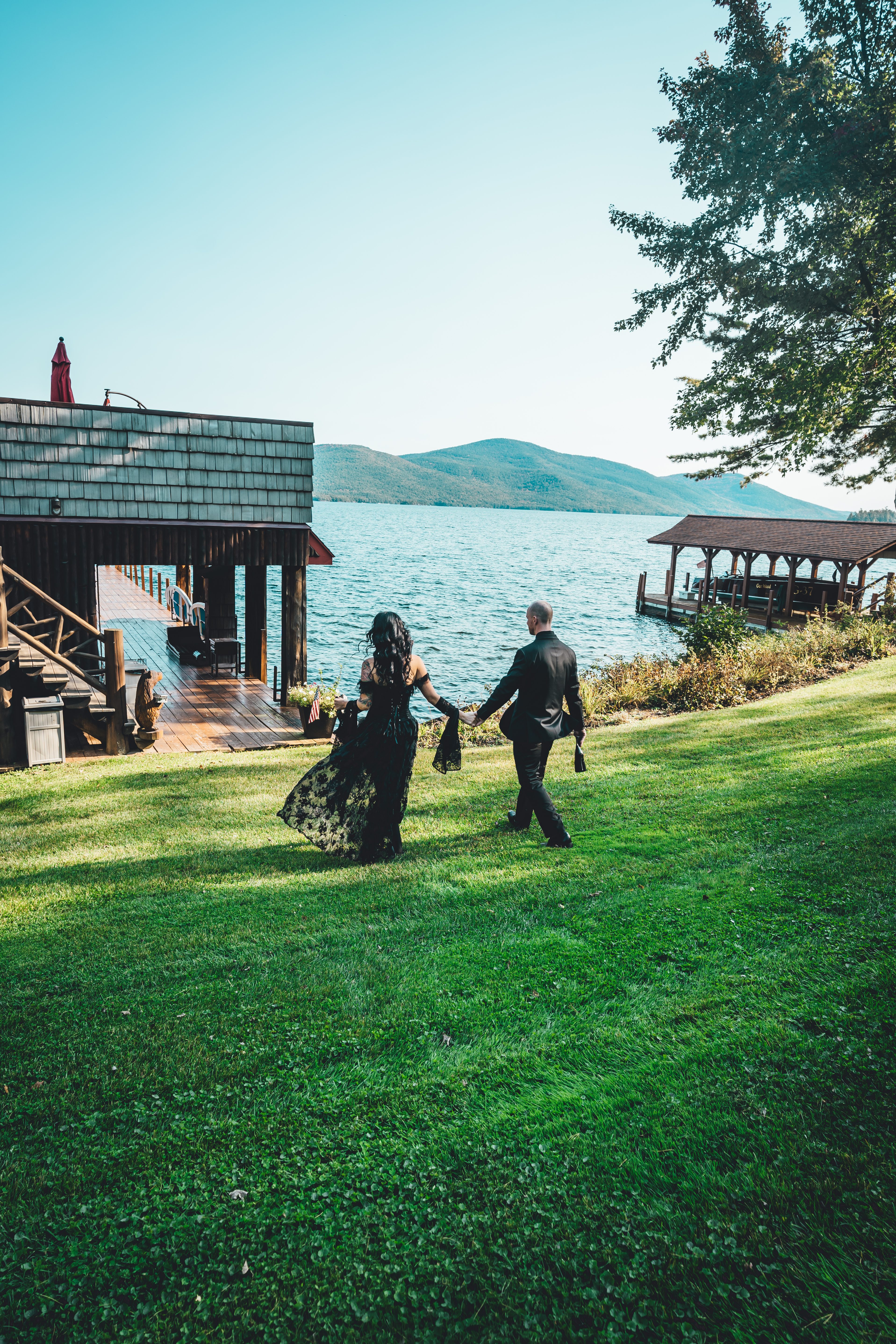 The bride in a black wedding dress and groom in black suit walk hand-in-hand across a manicured lawn toward a lakeside boathouse, with calm turquoise water and 