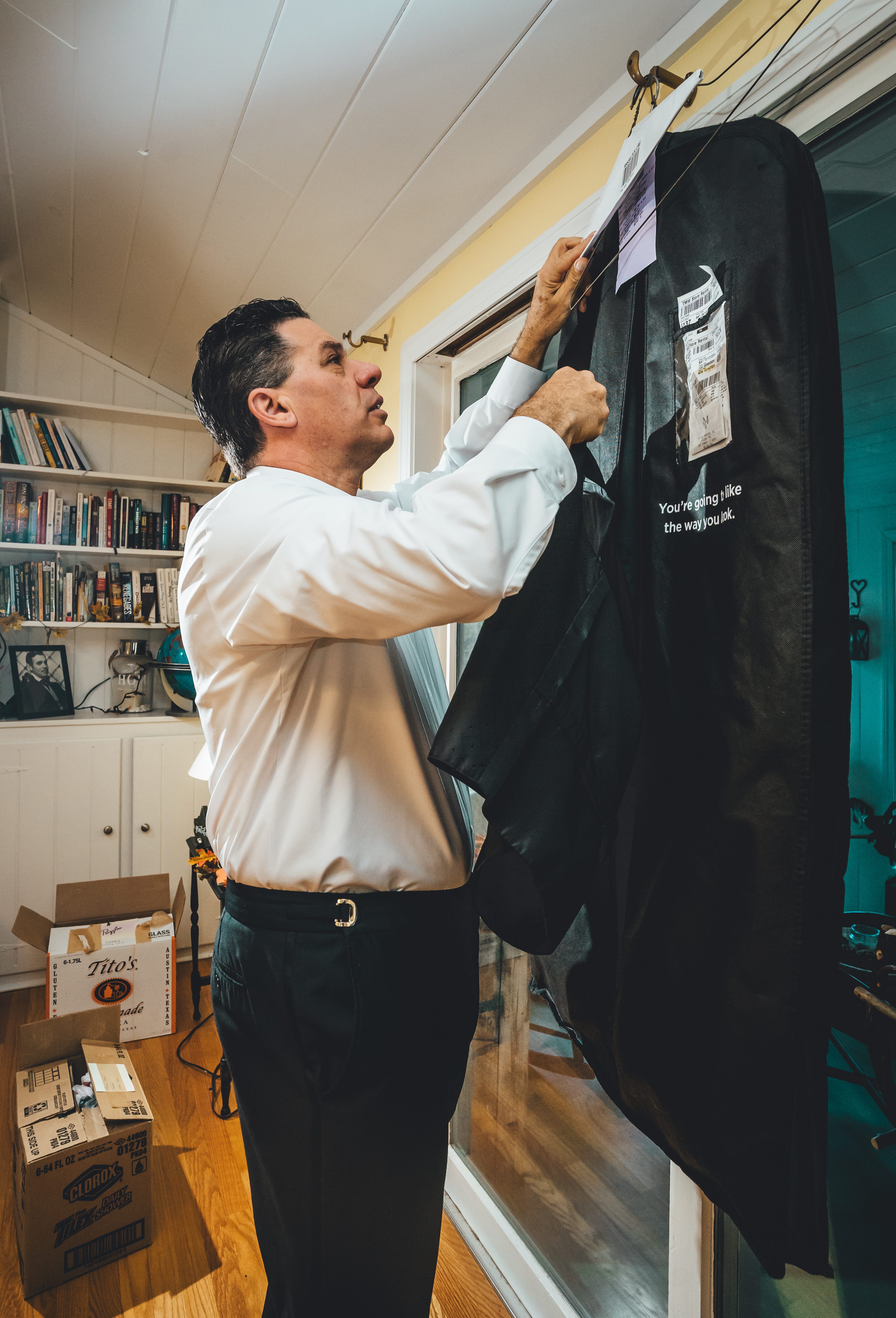 Groom Will carefully unpacks or examines his black wedding suit jacket from garment bag in a home office/library setting. Behind him are bookshelves filled with