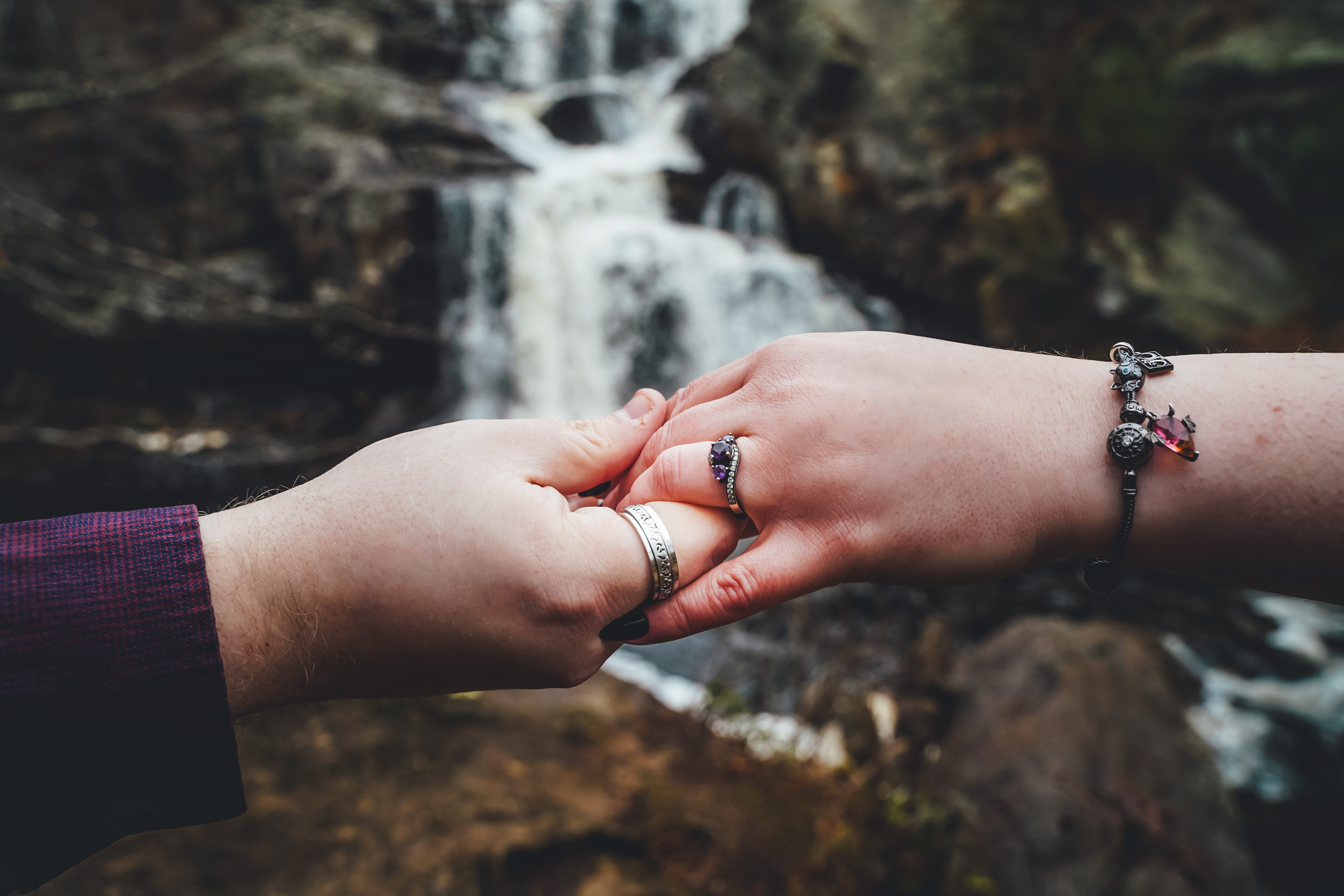 Close-up of two hands joined together displaying wedding bands in front of a cascading waterfall. The couple's hands are prominently featured with rings visible