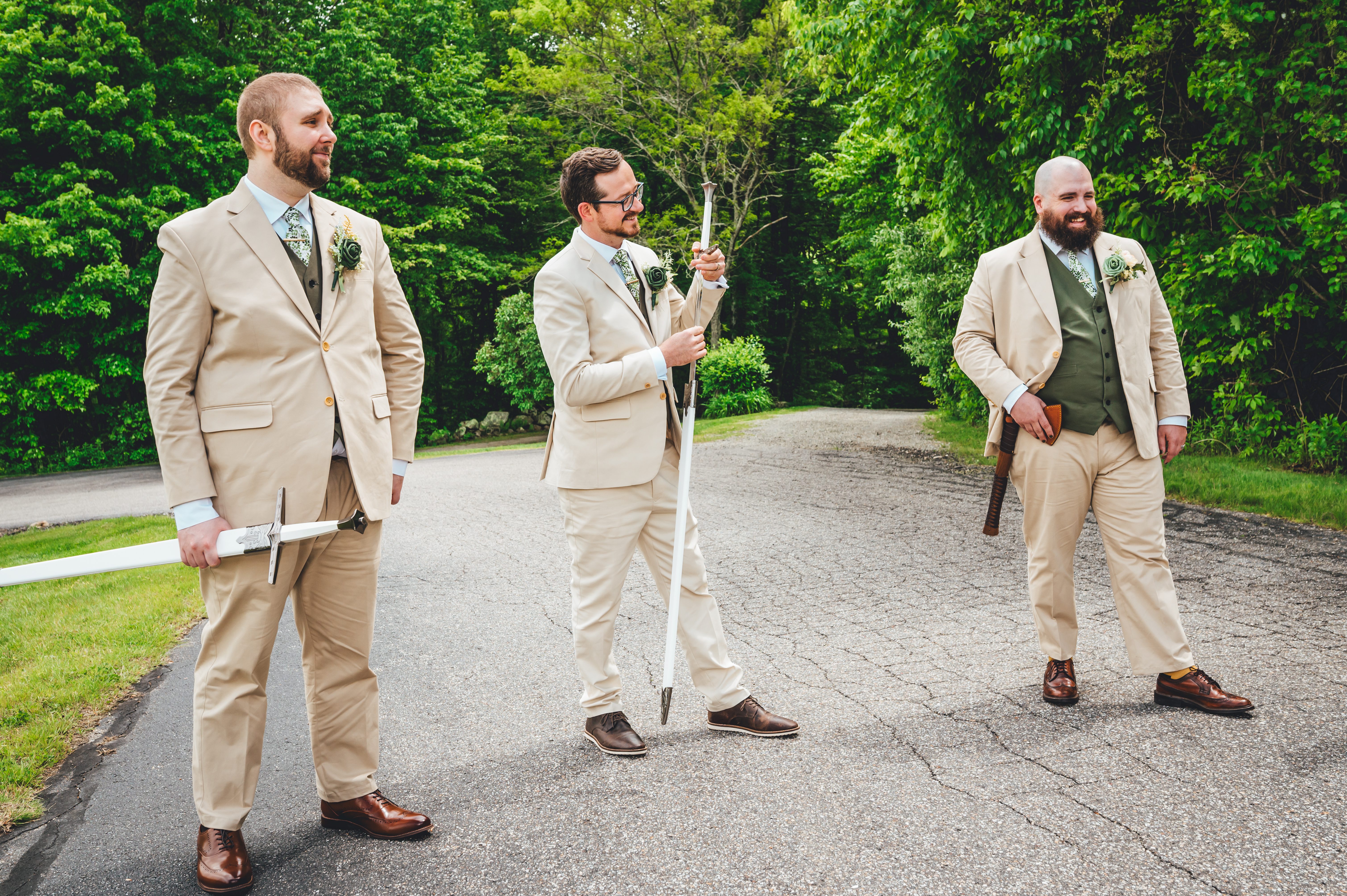 Three groomsmen in cream suits with boutonnières pose on a driveway holding fantasy weapons, a sword, staff, and axe, agai