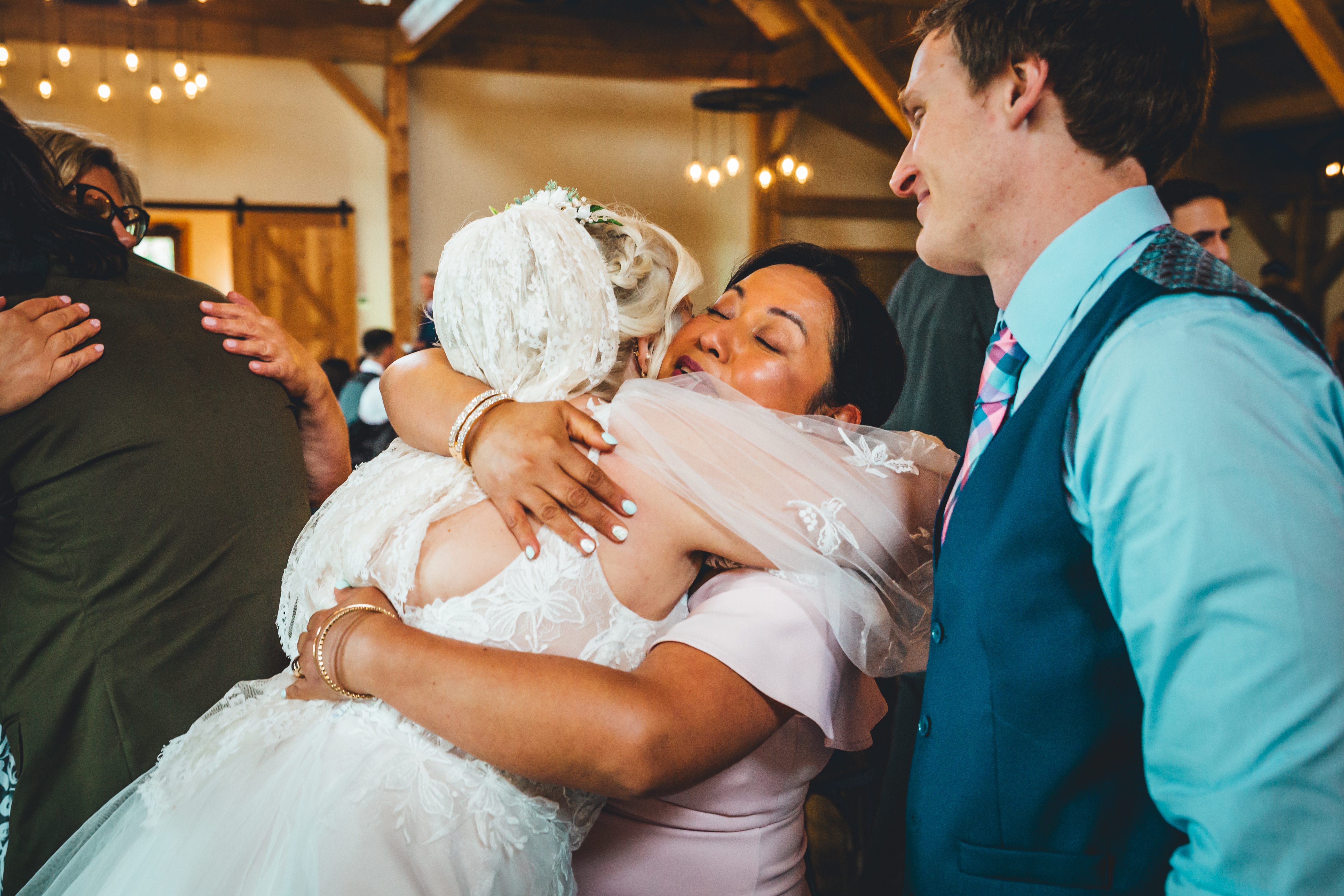 The bride in an elaborate white lace gown with an upswept hairstyle embraces a guest in a pink dress during the reception. The groom in a turquoise suit and pat