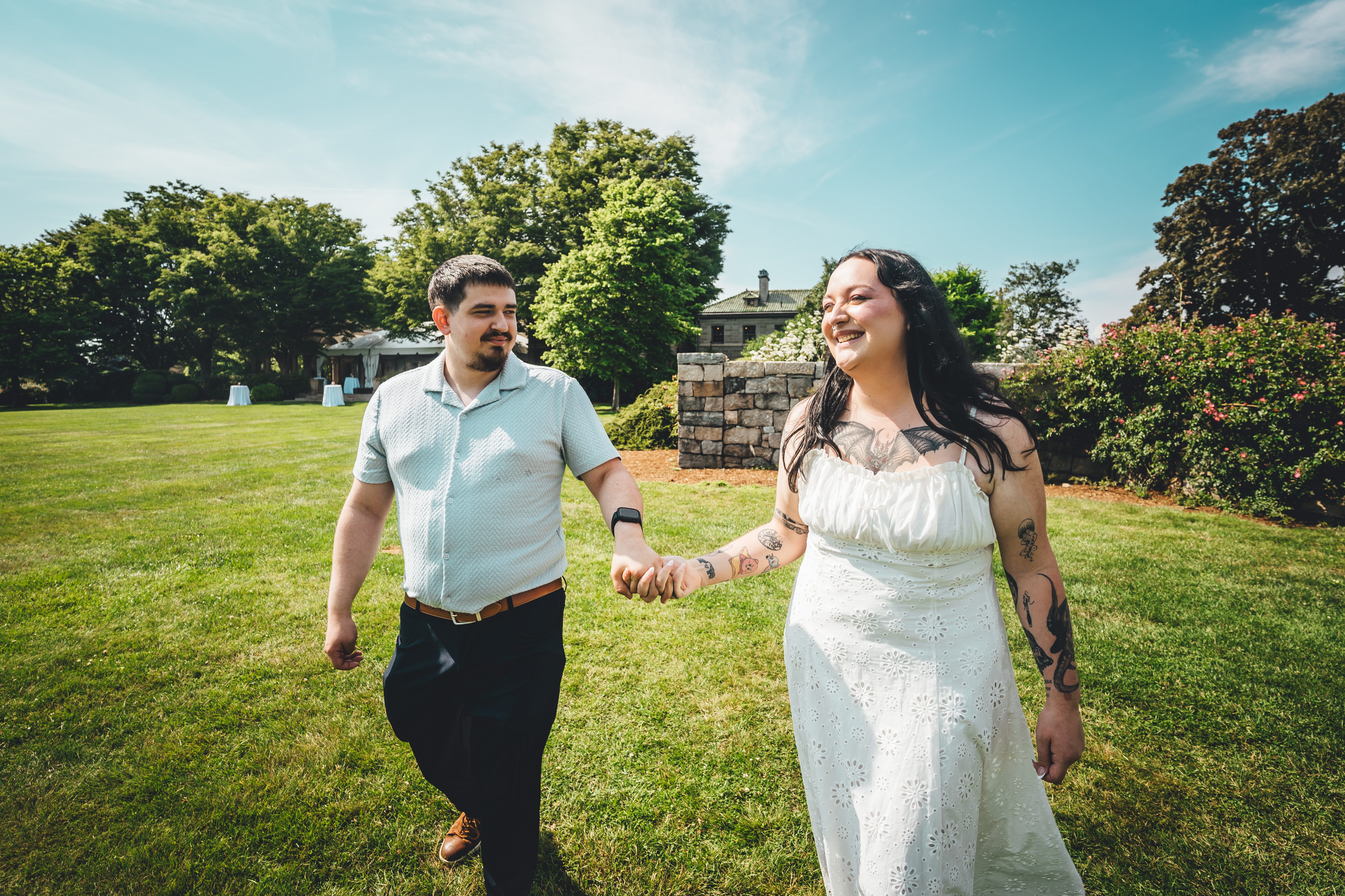 The bride and groom walk hand-in-hand across a lush green lawn on their wedding day. Both are smiling and looking toward each other, enjoying the moment. The br