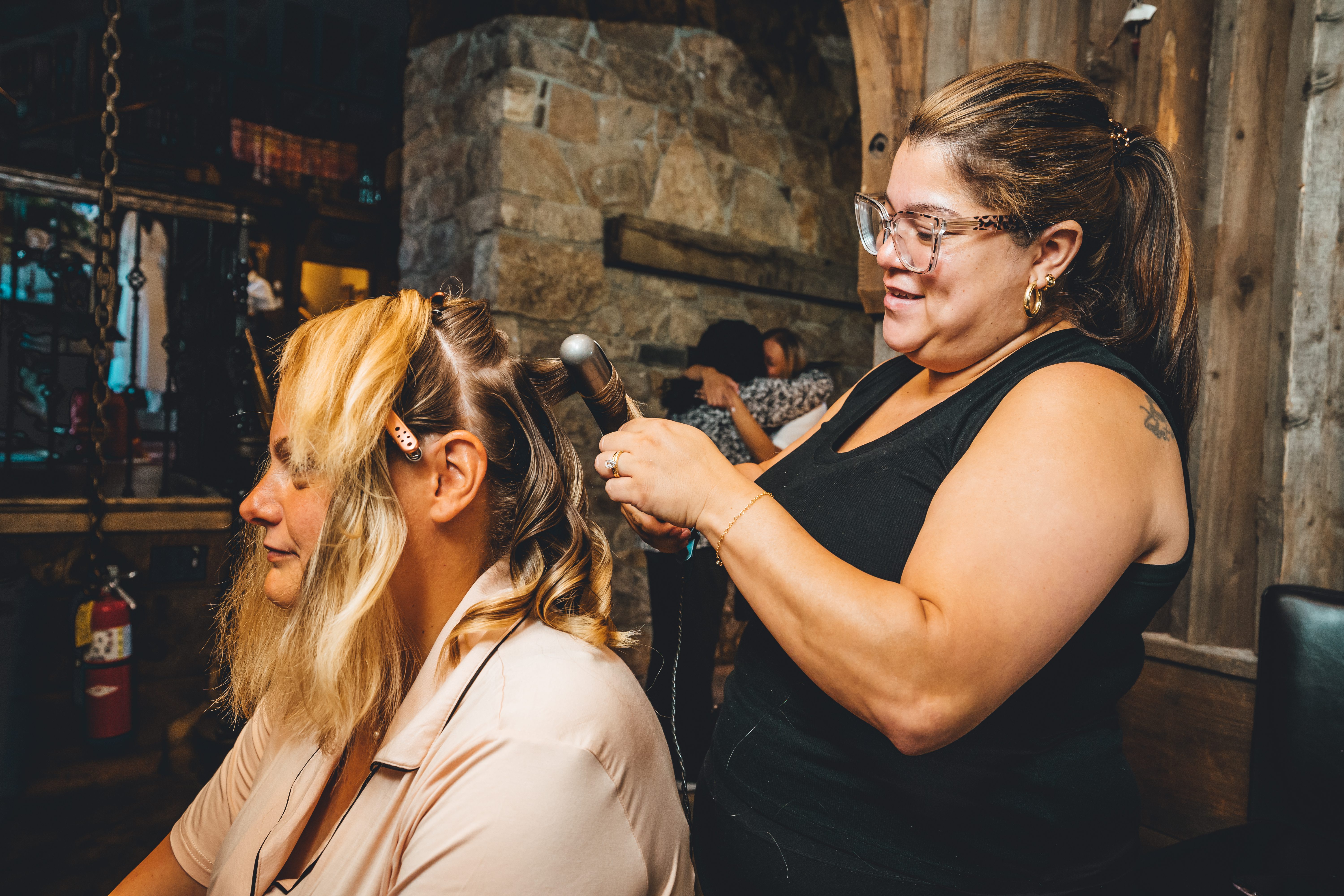 A stylist with glasses and dark hair blow-dries the bride's long blonde hair during wedding day preparations. The bride wears a soft beige/tan robe and sits sti