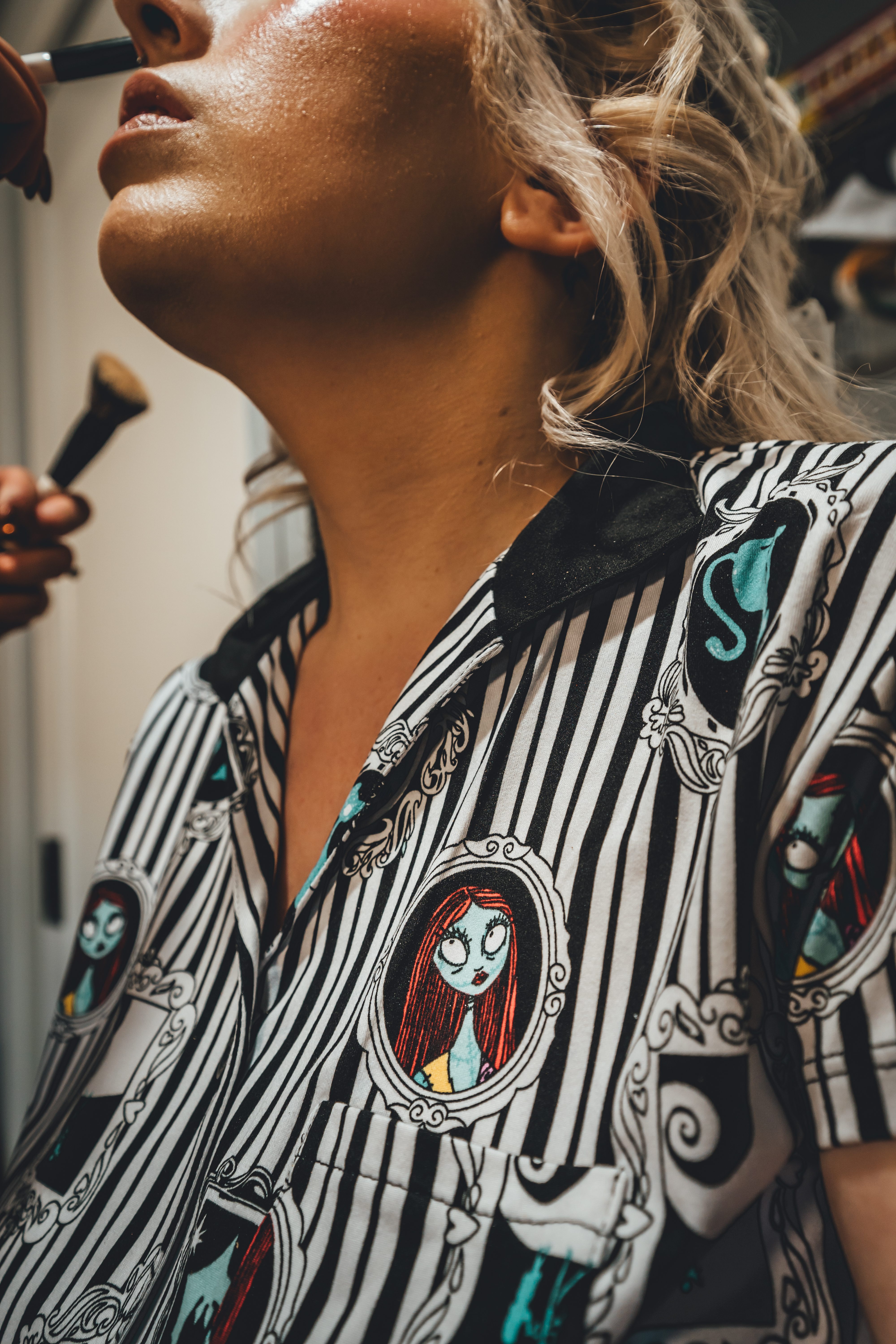 Close-up of a person's face and upper body during makeup application. They wear a striking black and white striped shirt featuring The Nightmare Before Christmas character Sally with ornate frame designs and gothic motifs in teal and red accents. A makeup brush is visible being applied near their face.