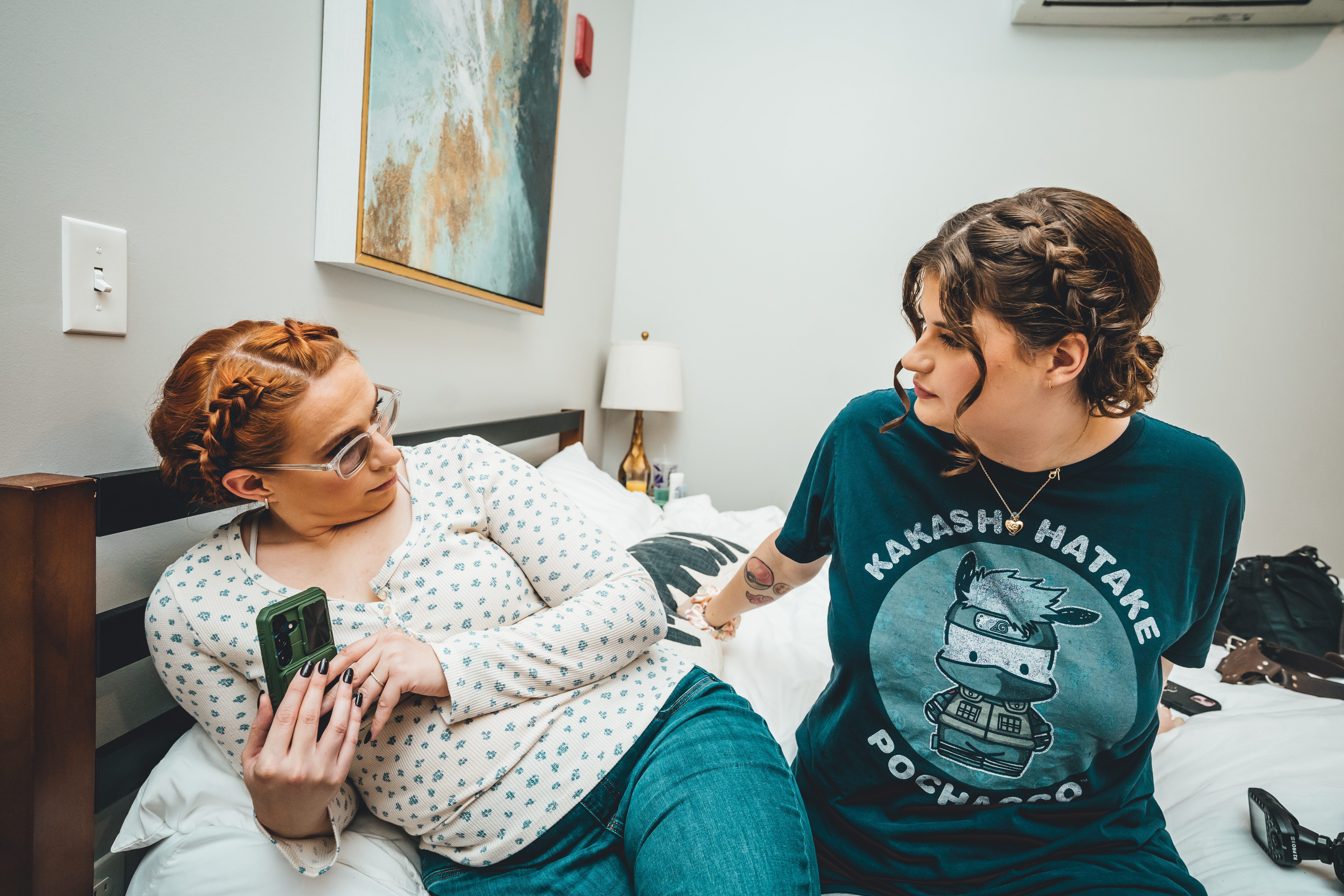 Two people relax on a bed in a hotel room during getting-ready, sitting close together and conversing. The person on the