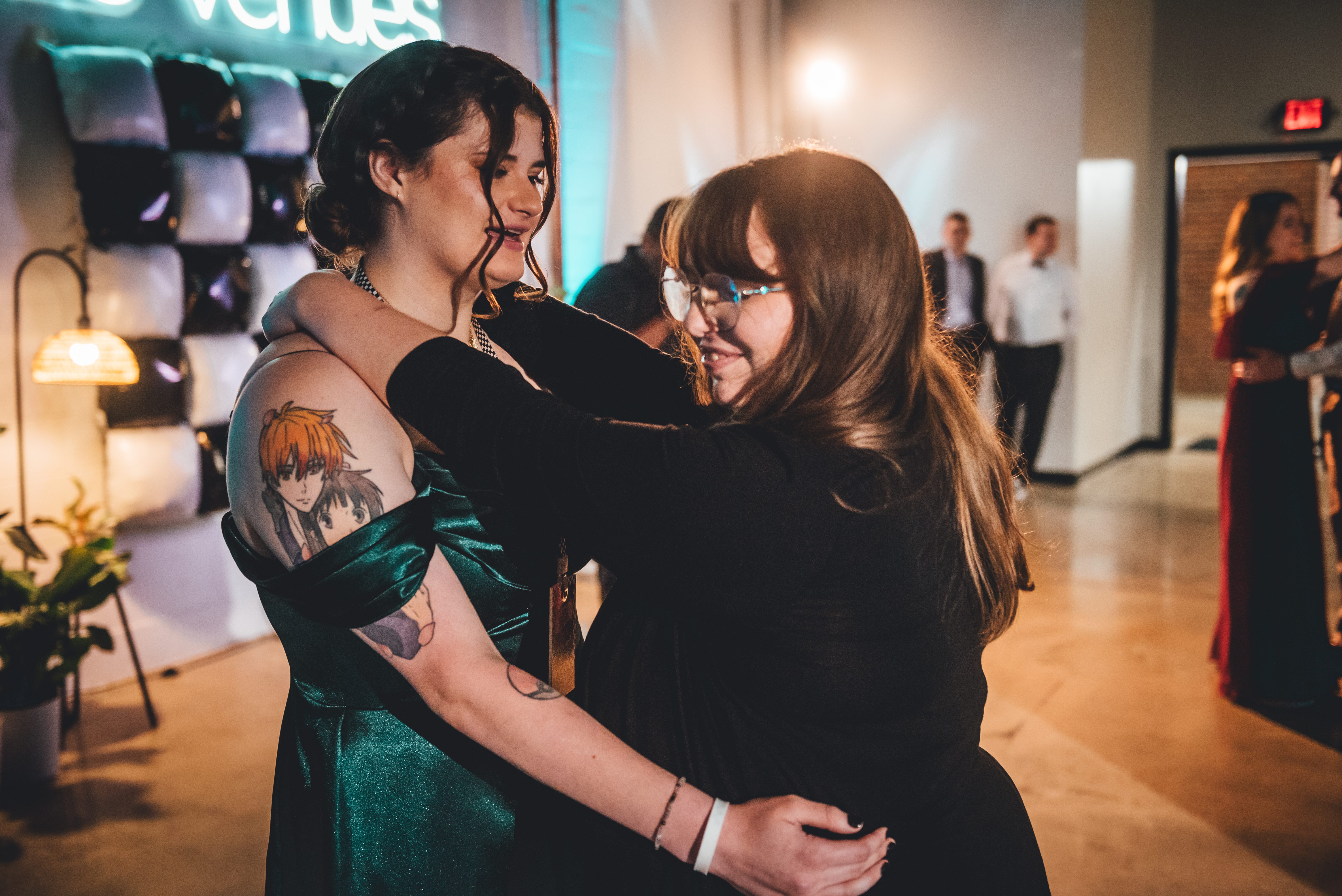 Two guests embrace warmly on the reception floor. The guest on the left wears a teal/emerald dress and displays an anime character tattoo on her shoulder, while