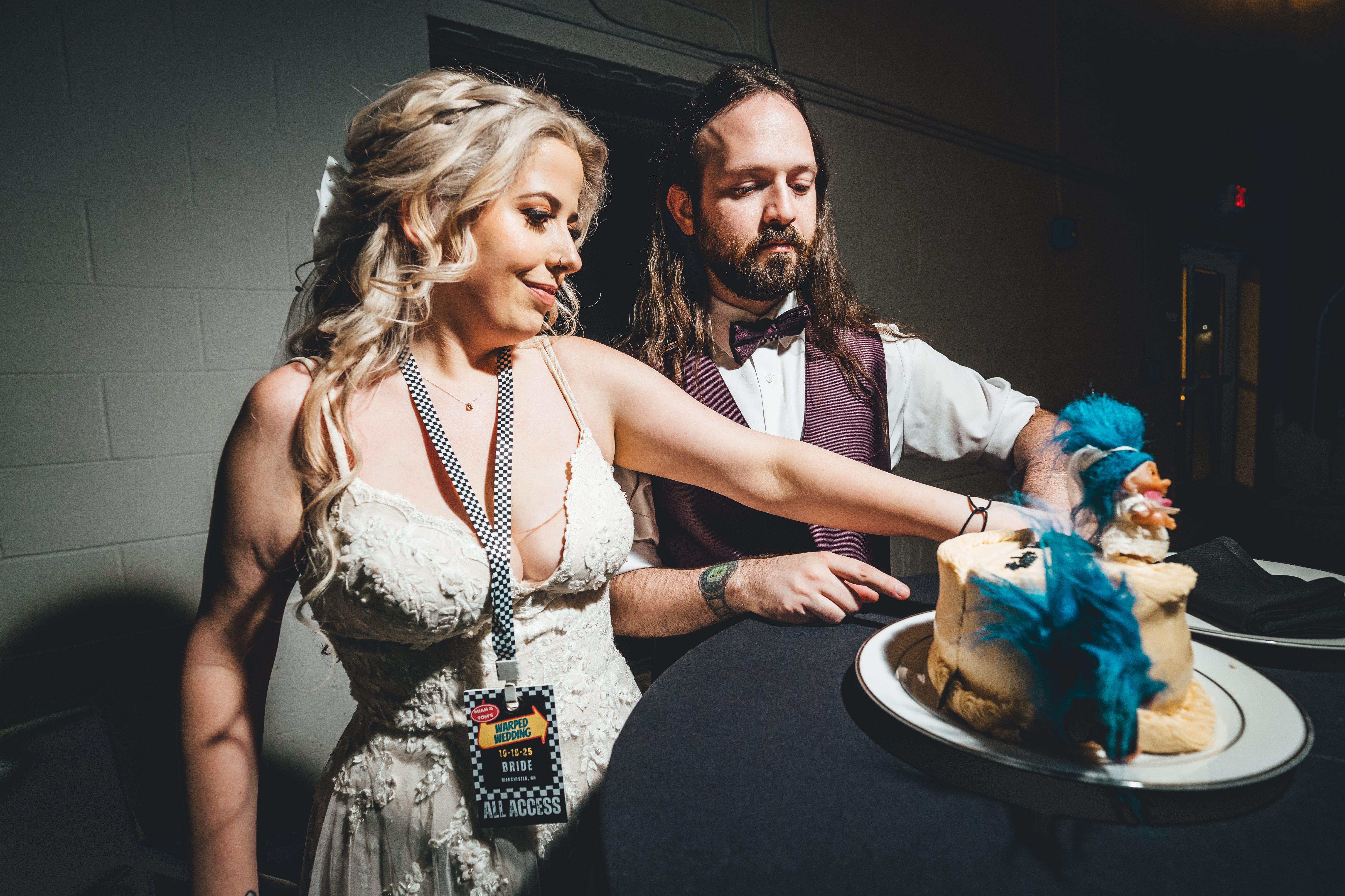 The bride and groom prepare to cut their wedding cake together indoors. The bride wears a white lace dress with a black-and-white checkered tie and an 'ALL ACCE