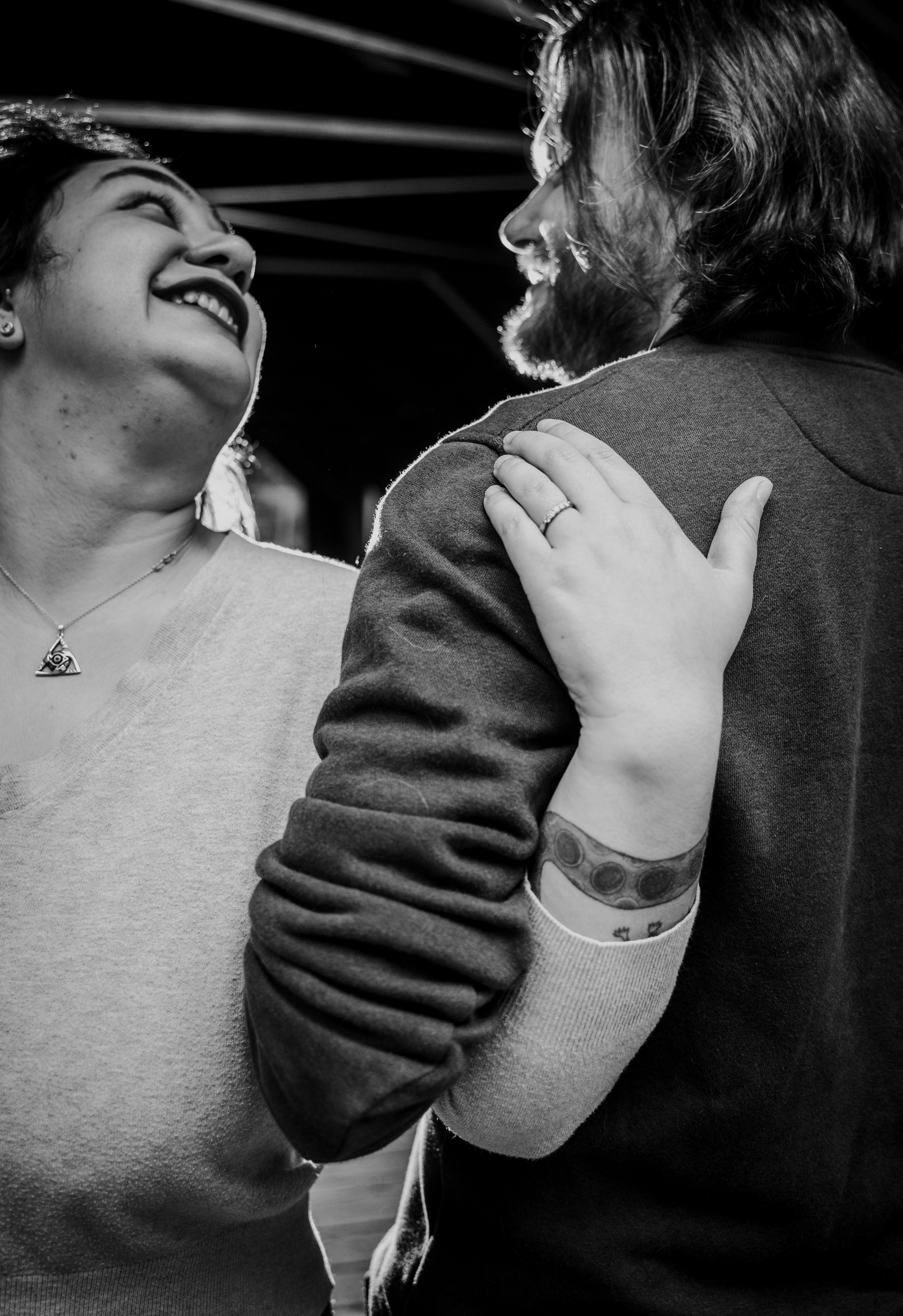 The couple embraces joyfully in close-up, both laughing and gazing at each other with genuine affection. The bride wears a triangular pendant necklace and displ