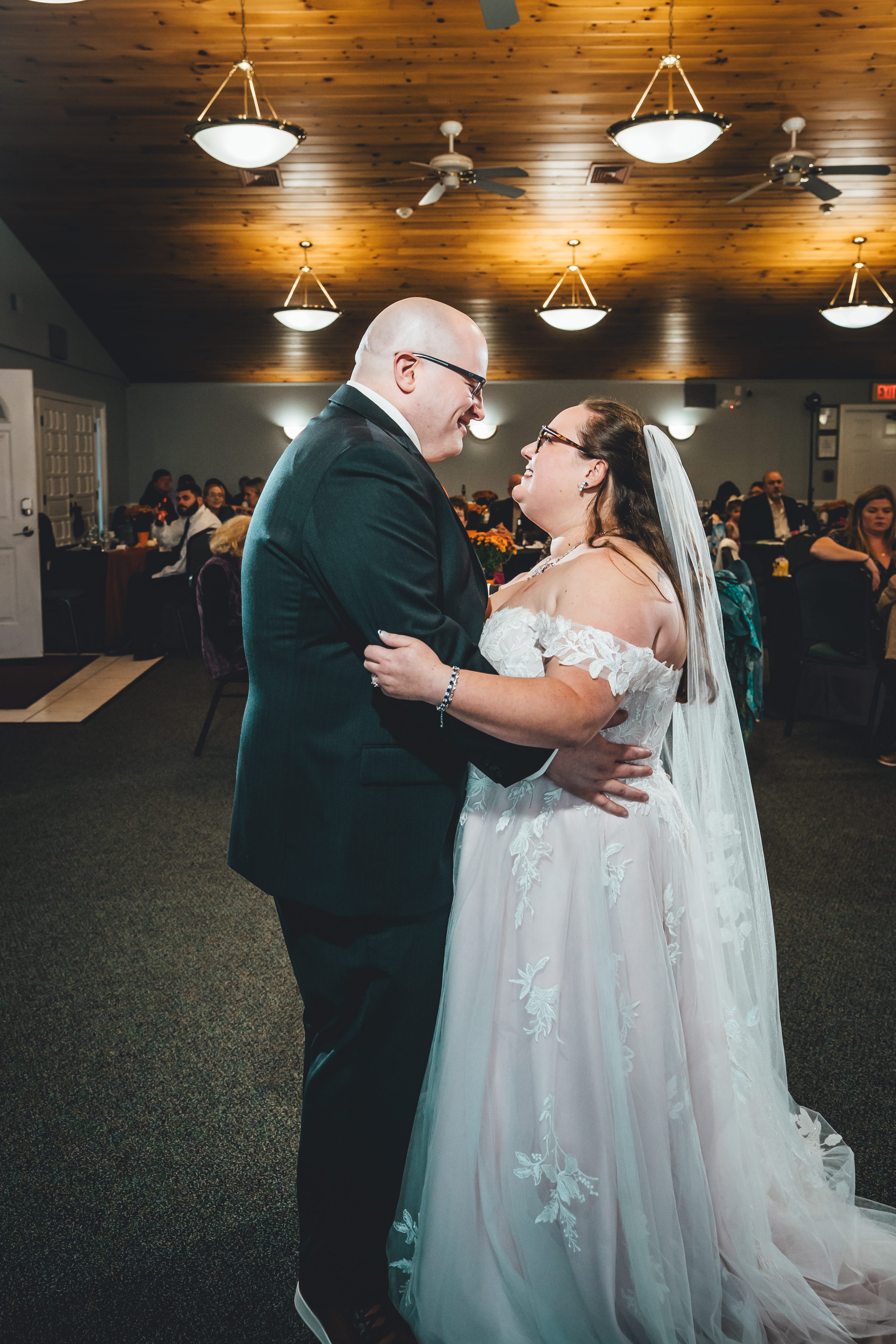 The couple shares an intimate moment on the reception floor, facing each other with joyful expressions. The groom wears a dark teal suit while the bride is in a