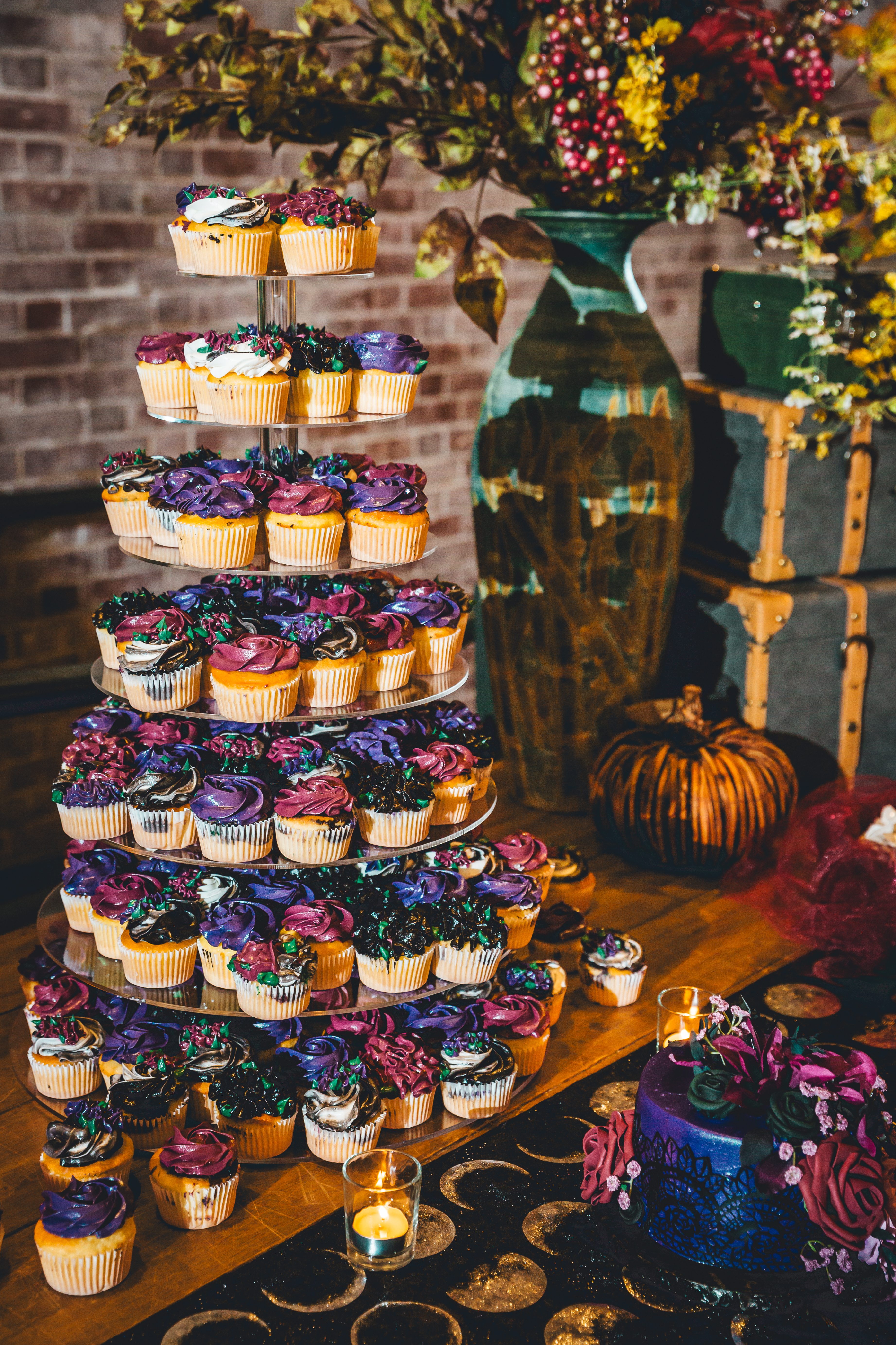 A towering tiered cupcake display with vibrant purple, pink, and blue frosting decorations stands against exposed brick. Behind it sits a vintage suitcase decor