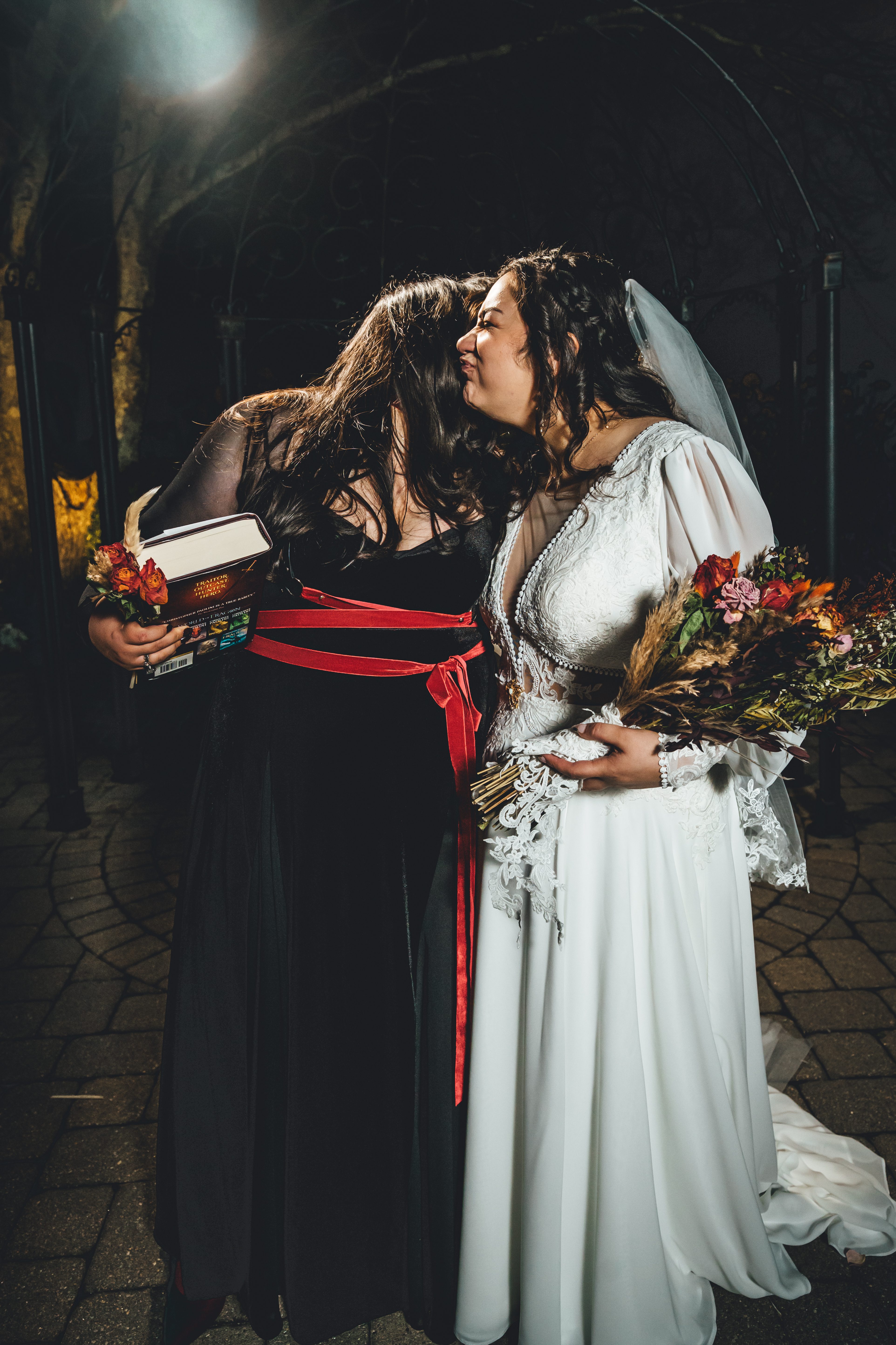 Two brides embrace under a stone archway at night, one wearing a white lace gown with veil holding a Stephen King book, the other in black with a red sash. Both