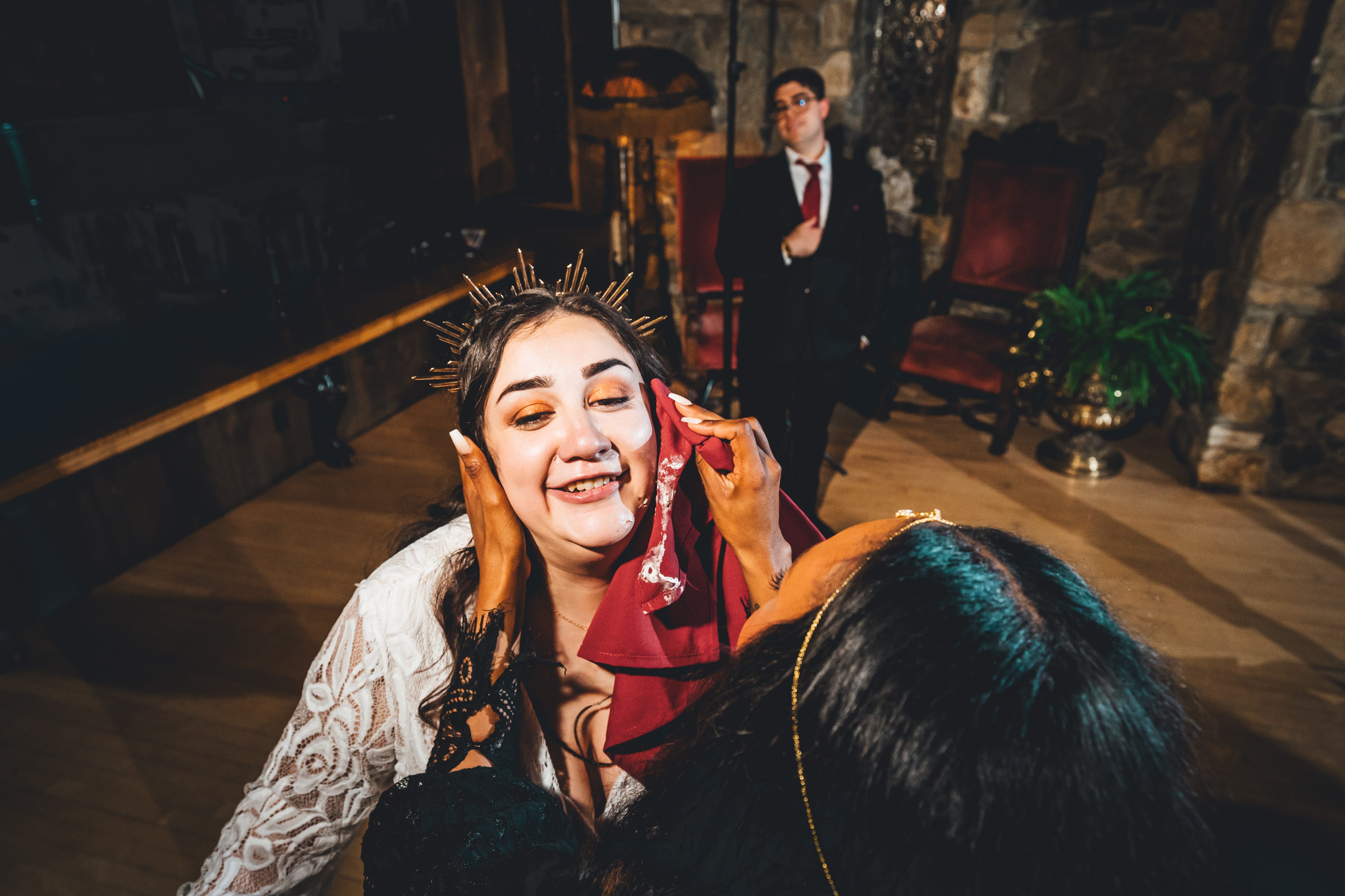 Bride wearing a dramatic spiked crown and bold orange eyeshadow laughs joyfully while holding a red fabric with a guest during reception festivities. The groom 