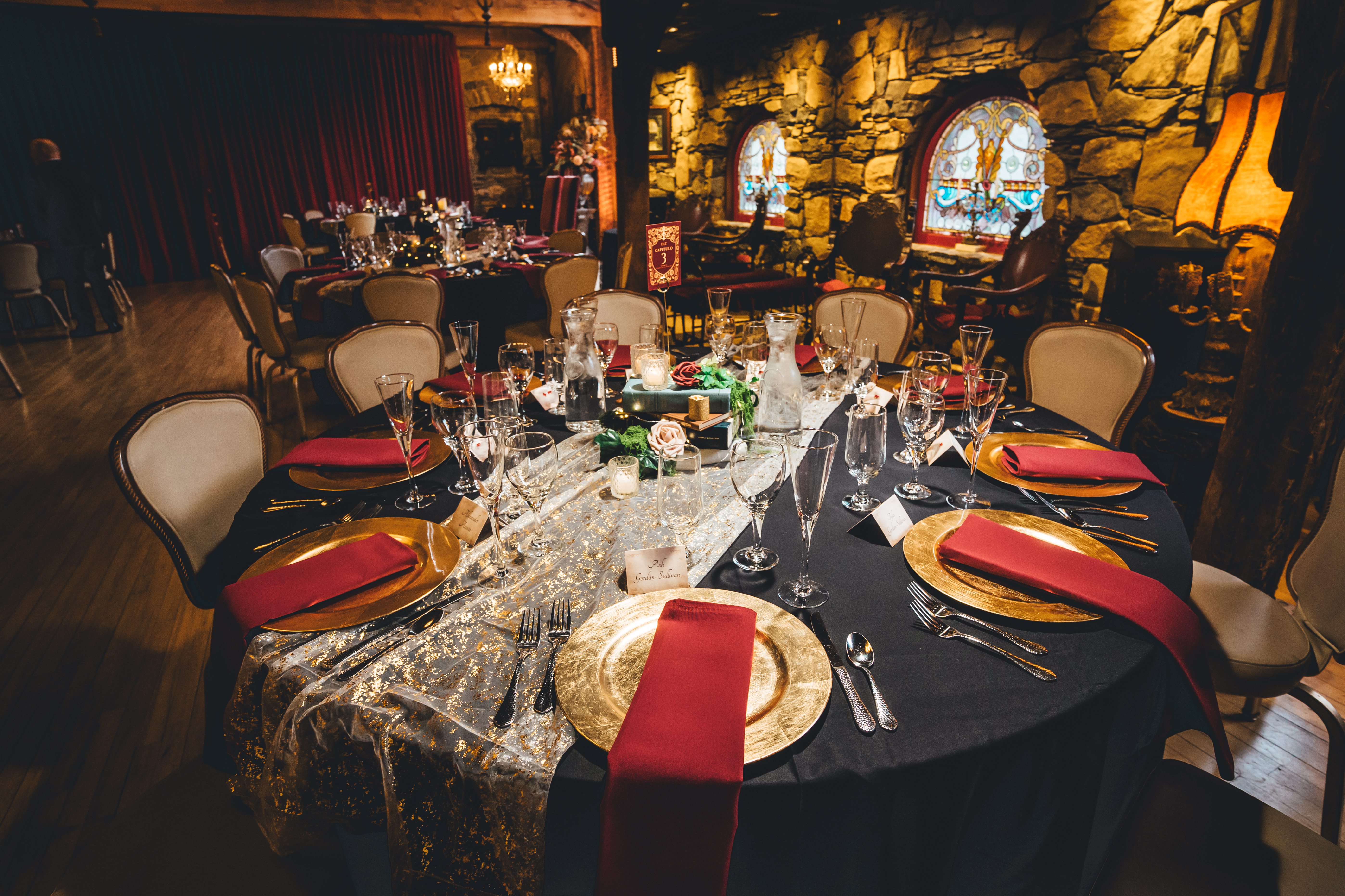An elegantly decorated reception table with black linens, gold charger plates, red napkins, and a gold-patterned table runner. The venue features dramatic stone