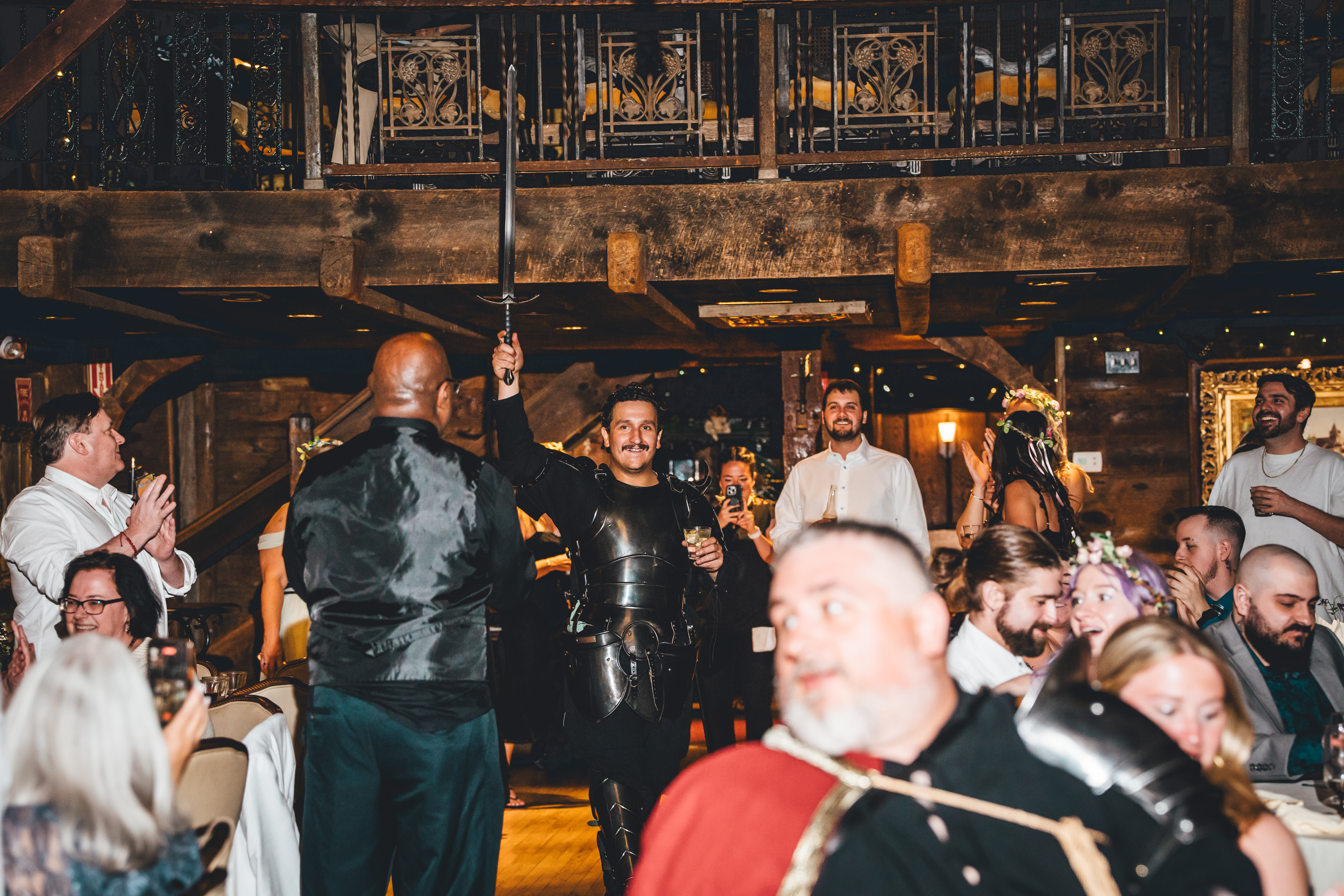A lively reception moment with guests gathered around the dance floor in a rustic venue. A man in the center wears full black armor cosplay, holding a sword alo