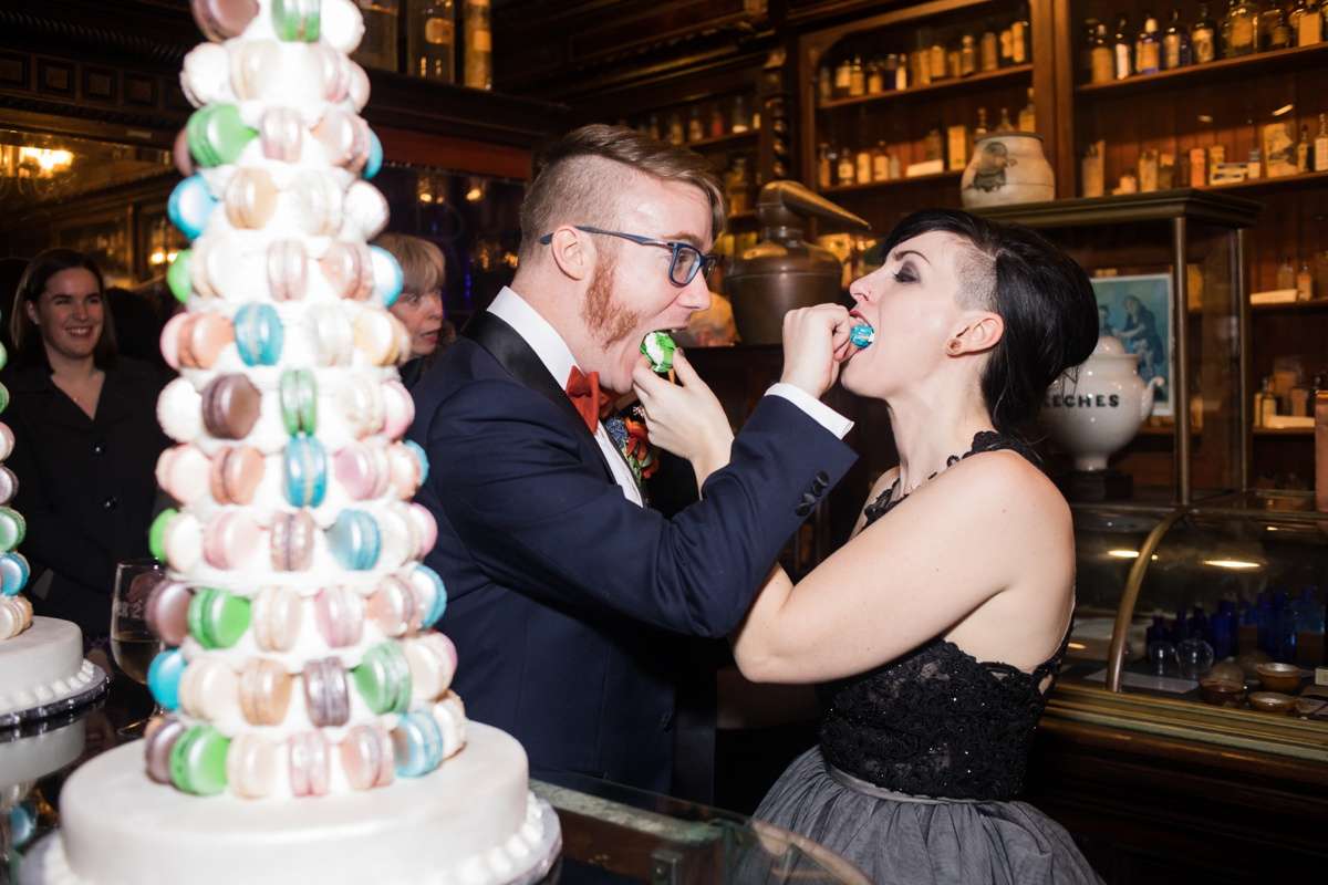 A couple feeds each other macarons beside their macaron tower wedding cakes