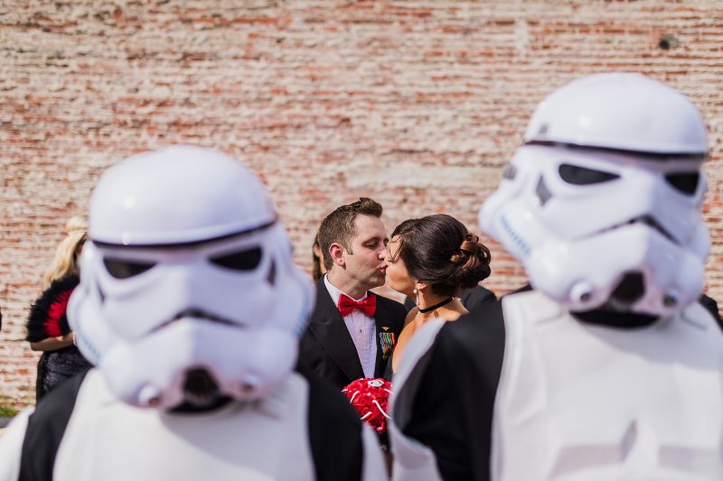 A couple kisses framed between two stormtrooper helmets at their May the Fourth wedding