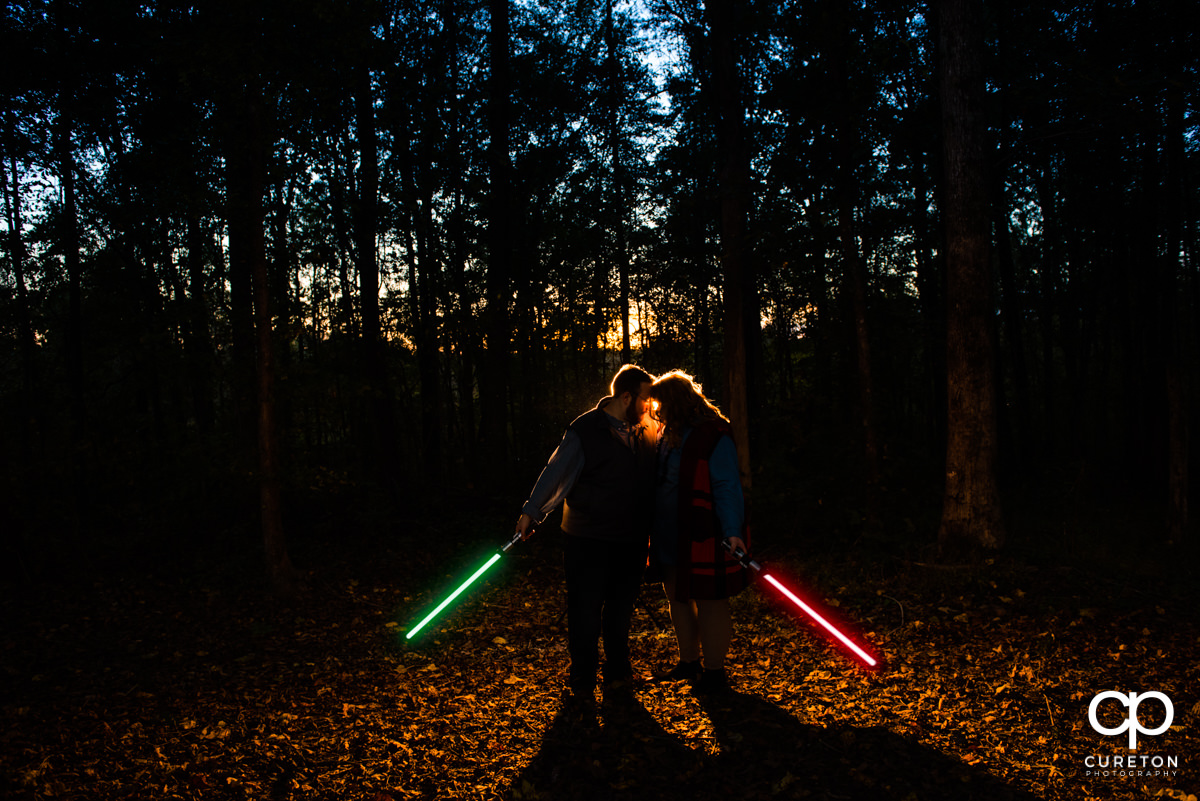 An engaged couple holds green and red lightsabers in a forest at sunset